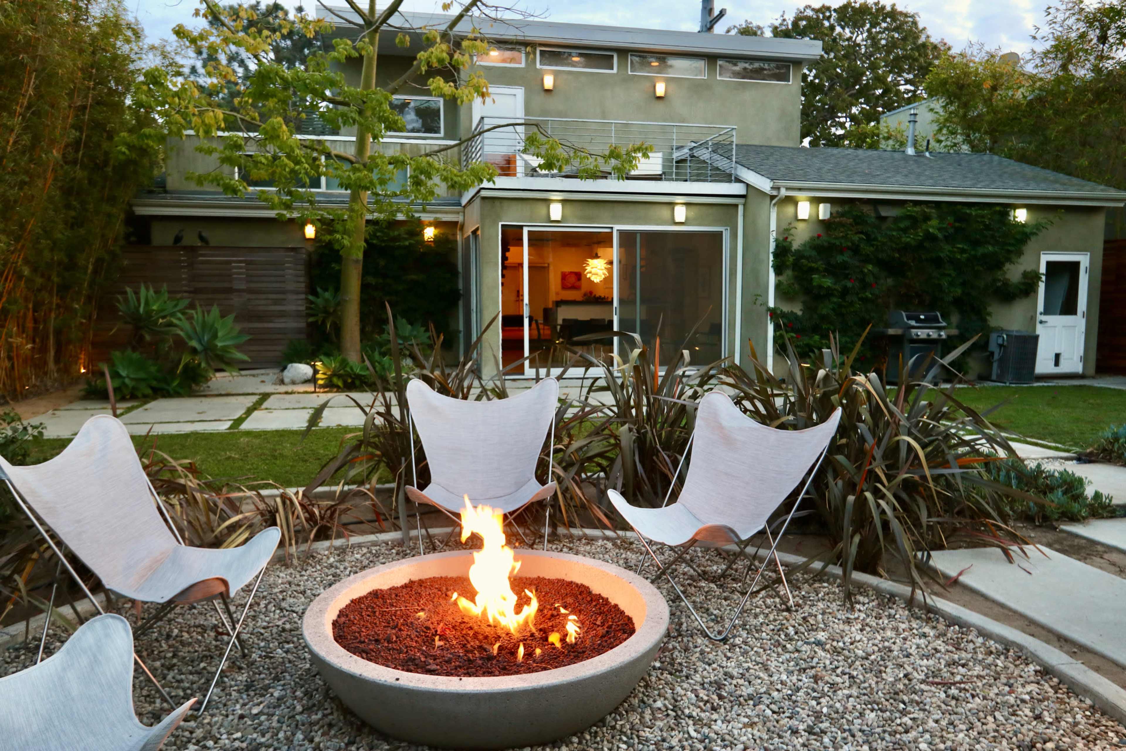 A modern backyard features a circular fire pit surrounded by four butterfly chairs, with a house and lush greenery in the background.