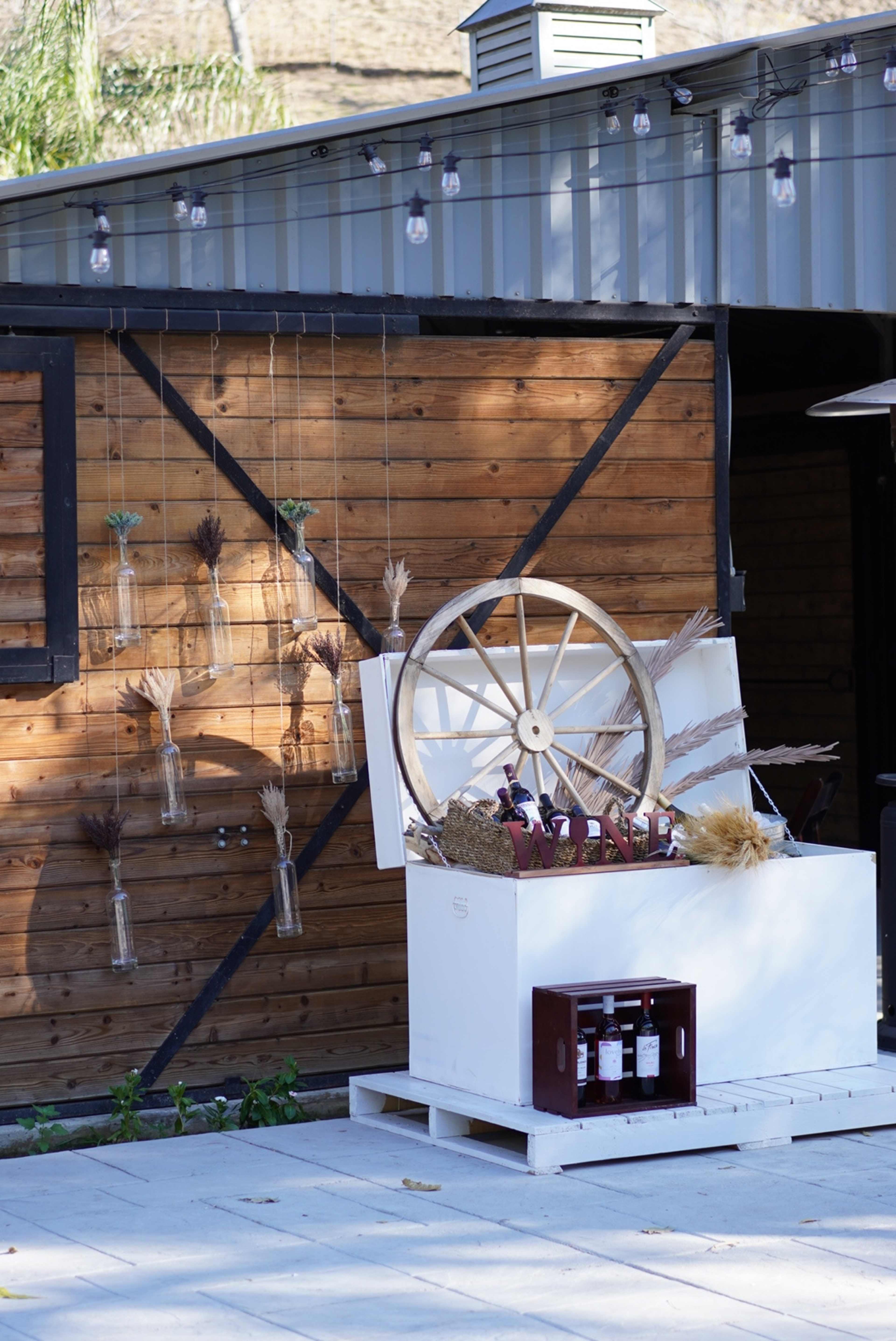 A rustic wooden structure features a decorative display with a large wagon wheel and hanging glass vases, alongside a white crate holding several bottles of wine.