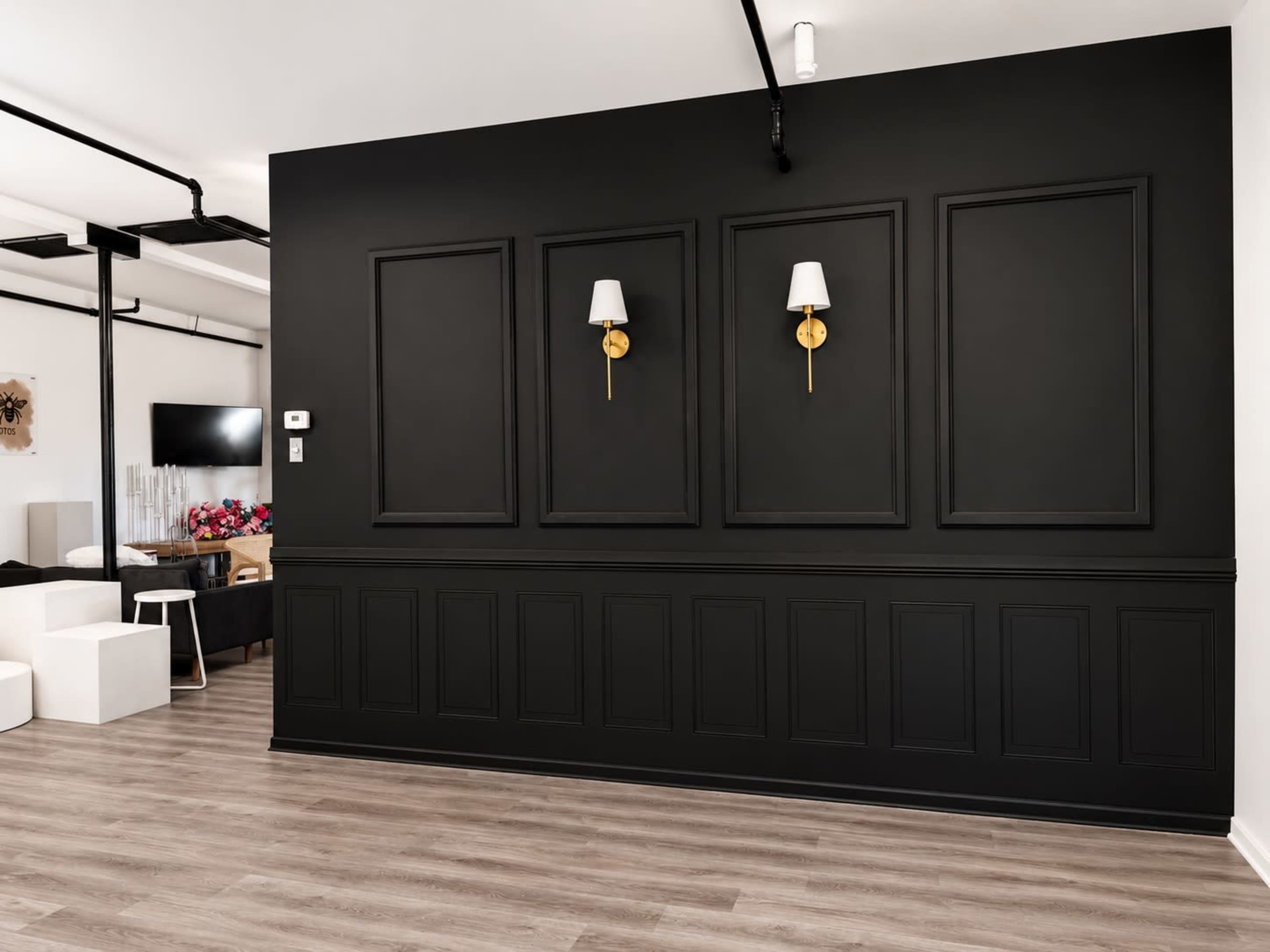 The image shows a modern room with a black wall featuring three framed sections and two wall sconces, alongside a light-colored floor and visible furnishings in the background.