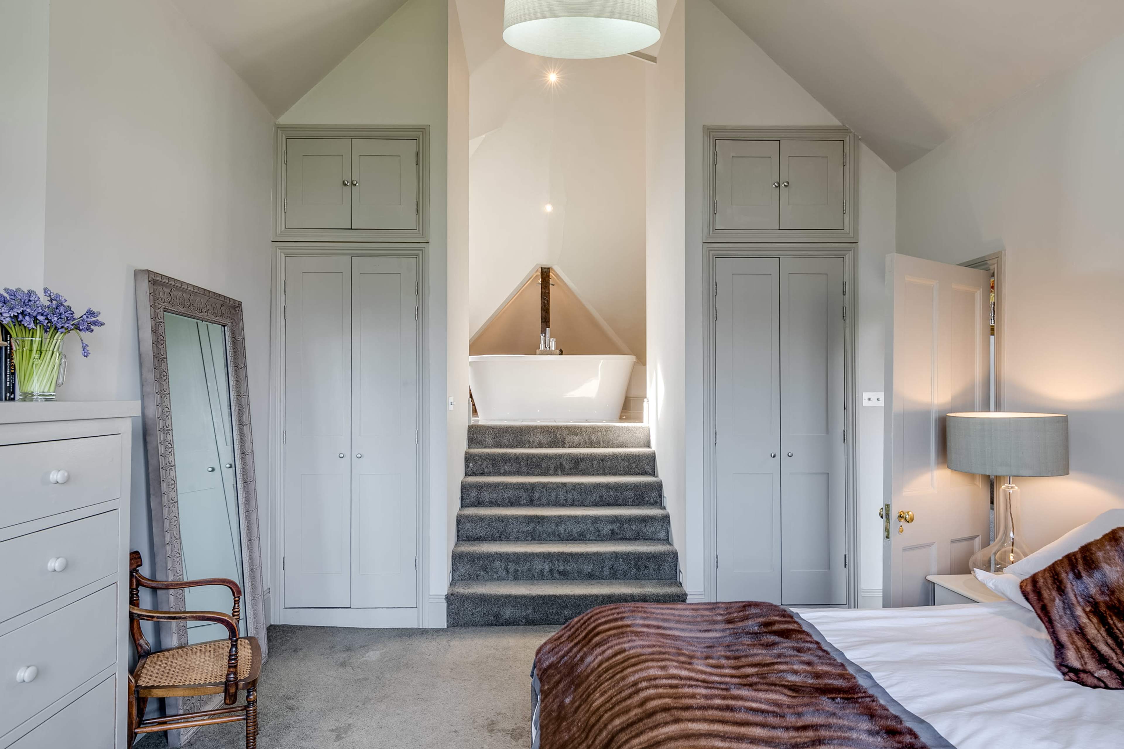 The image shows a minimalist bedroom with a bed, a staircase leading to a bathtub, grey cabinets flanking the entrance, and a chair beside a dresser.