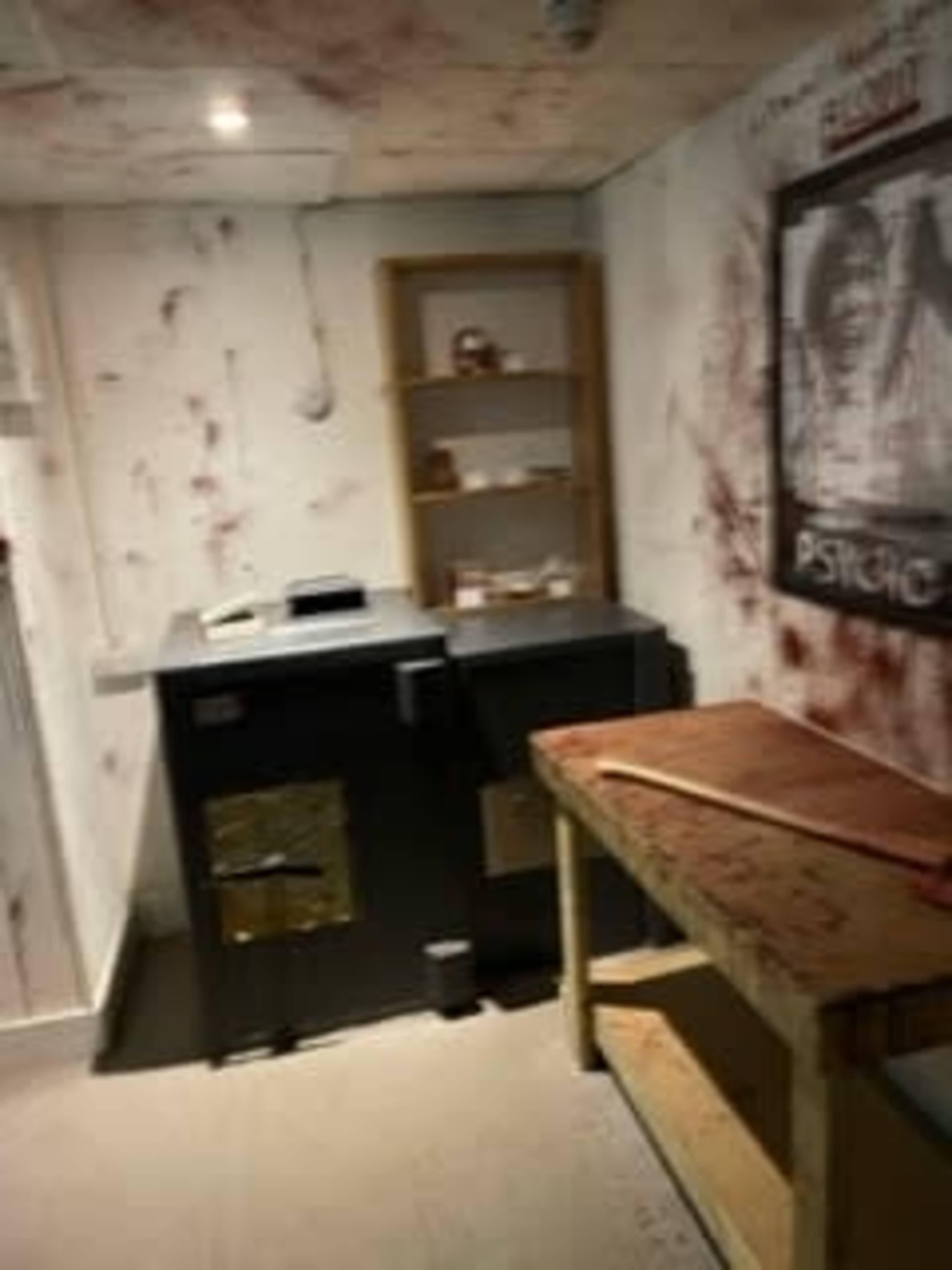 A dimly lit room with bloodstains on the walls, a safe on the left, a wooden table in the center, and a shelf with items in the background.