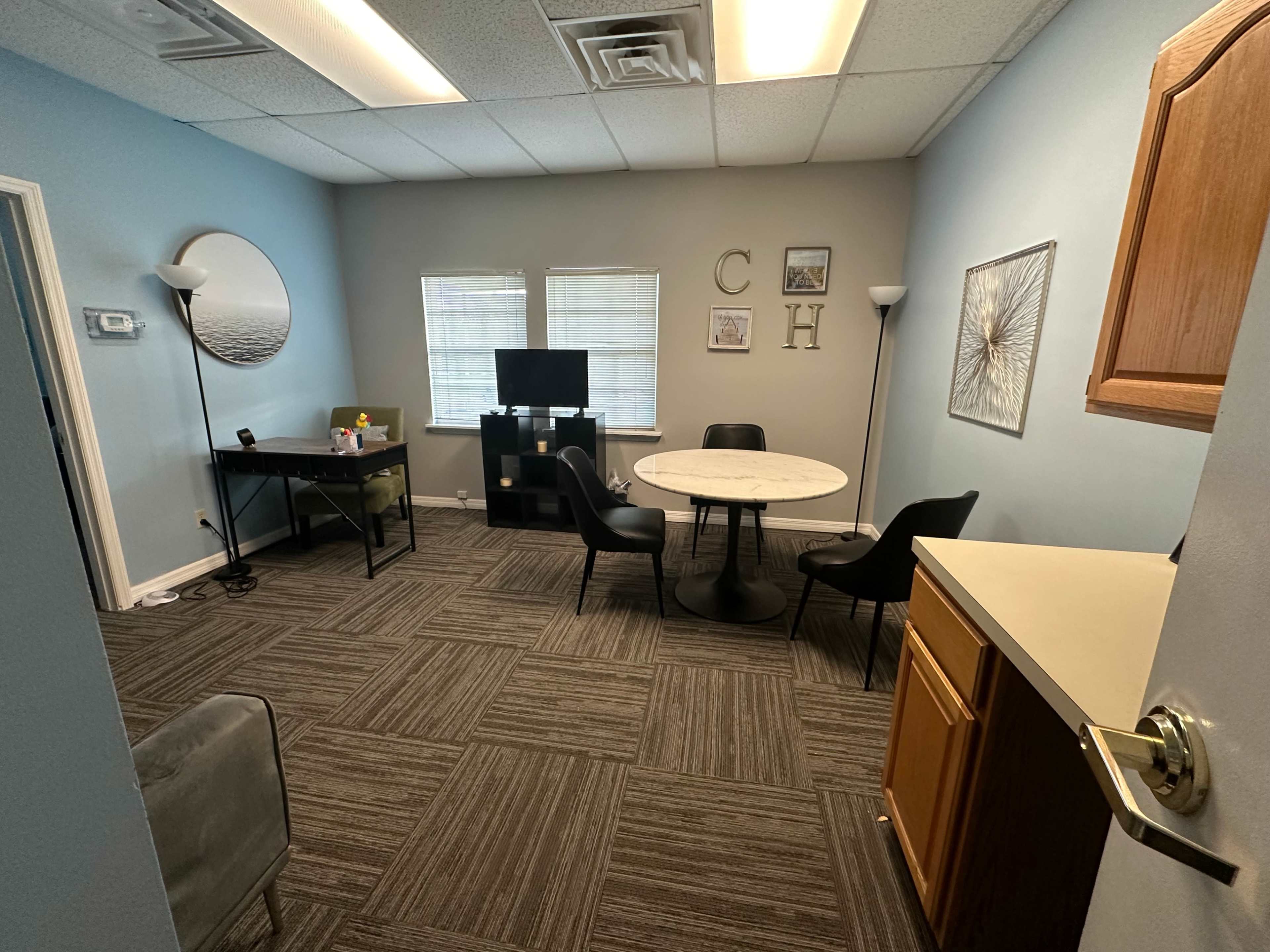 The image shows a small office space with a circular table, four chairs, a desk, a TV, and light blue walls.
