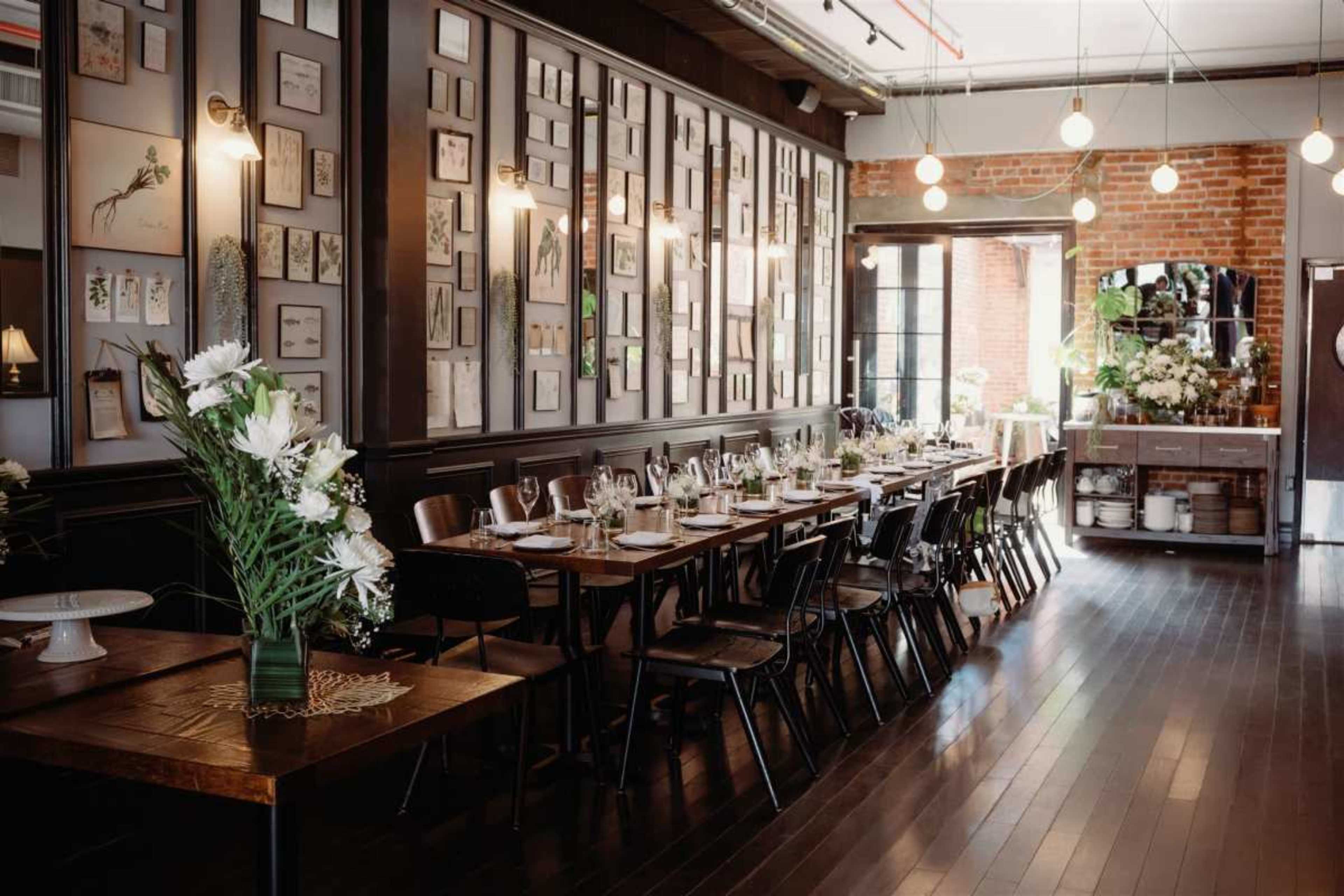 The image shows a spacious restaurant interior featuring a long wooden dining table set with glassware, surrounded by black chairs, and adorned with framed artwork on the walls.