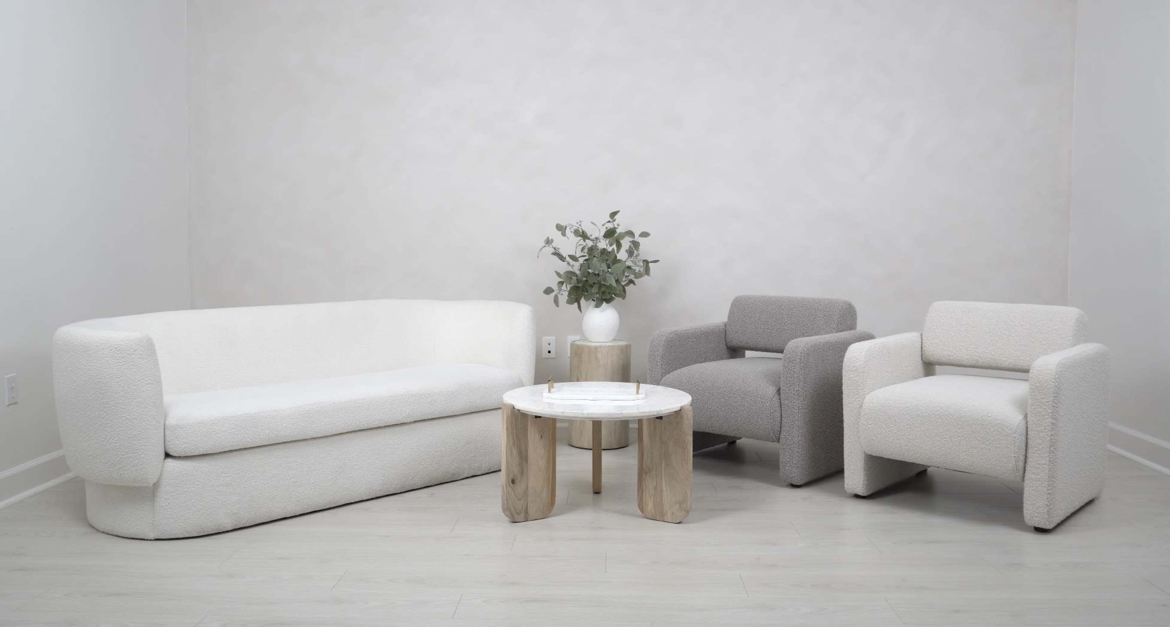 A modern living room setup features a white upholstered sofa, two accent chairs in light gray and white, and a round wooden coffee table adorned with a vase of greenery.