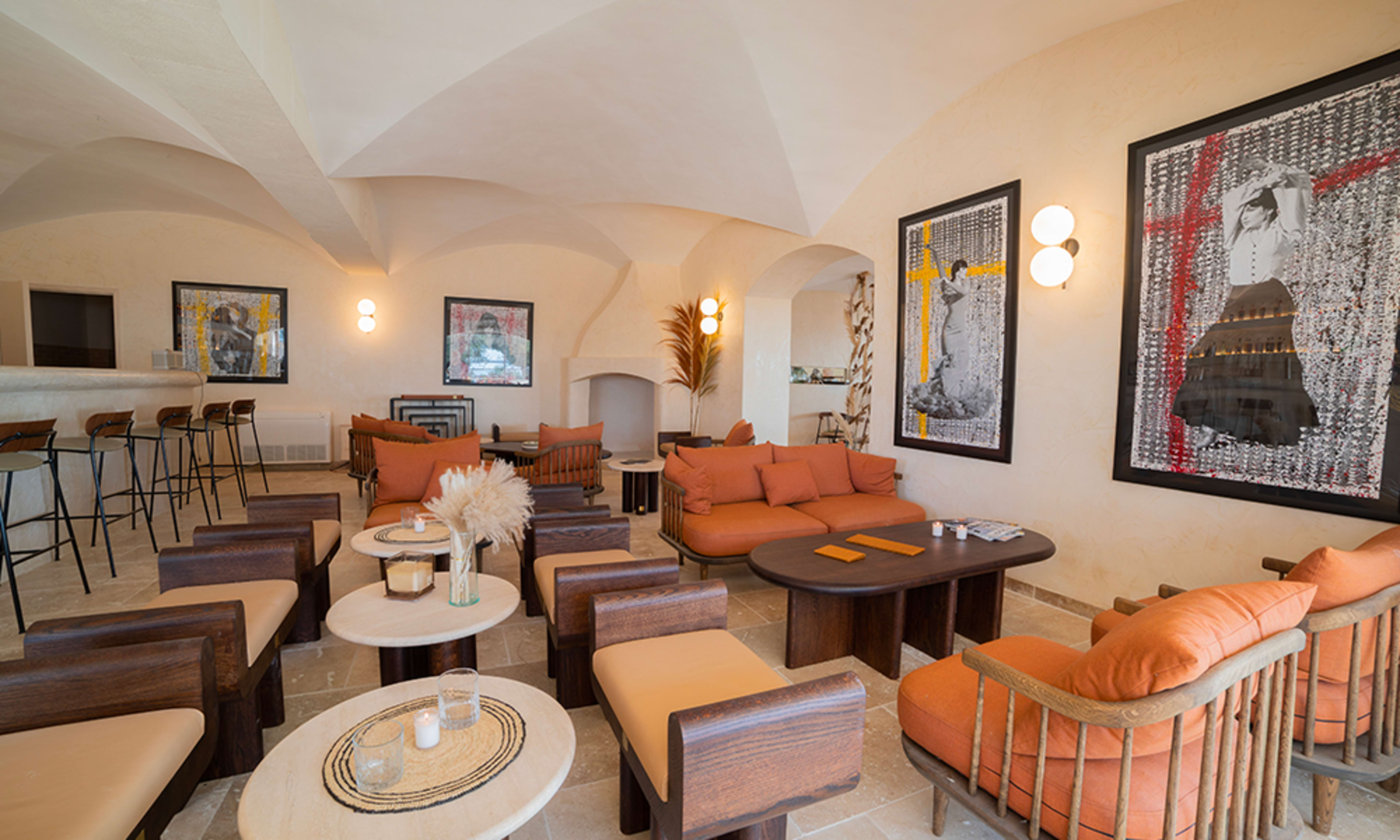 The image shows a modern, minimalist lounge area featuring orange and brown seating, round tables, and artwork on the walls.