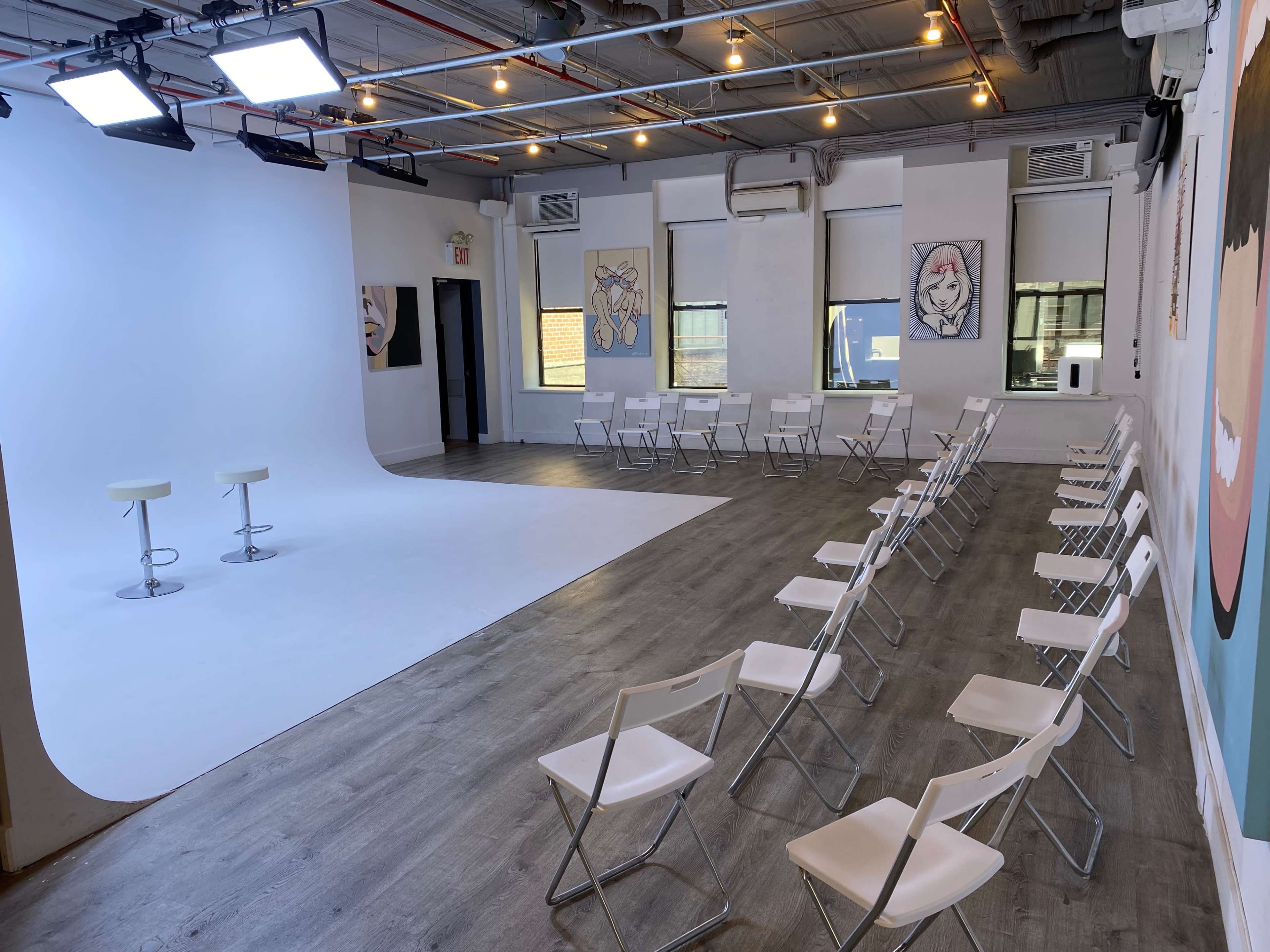 The image shows a spacious, well-lit room with white walls, featuring a curved backdrop, two stools on a stage-like area, and rows of white folding chairs arranged for an audience.