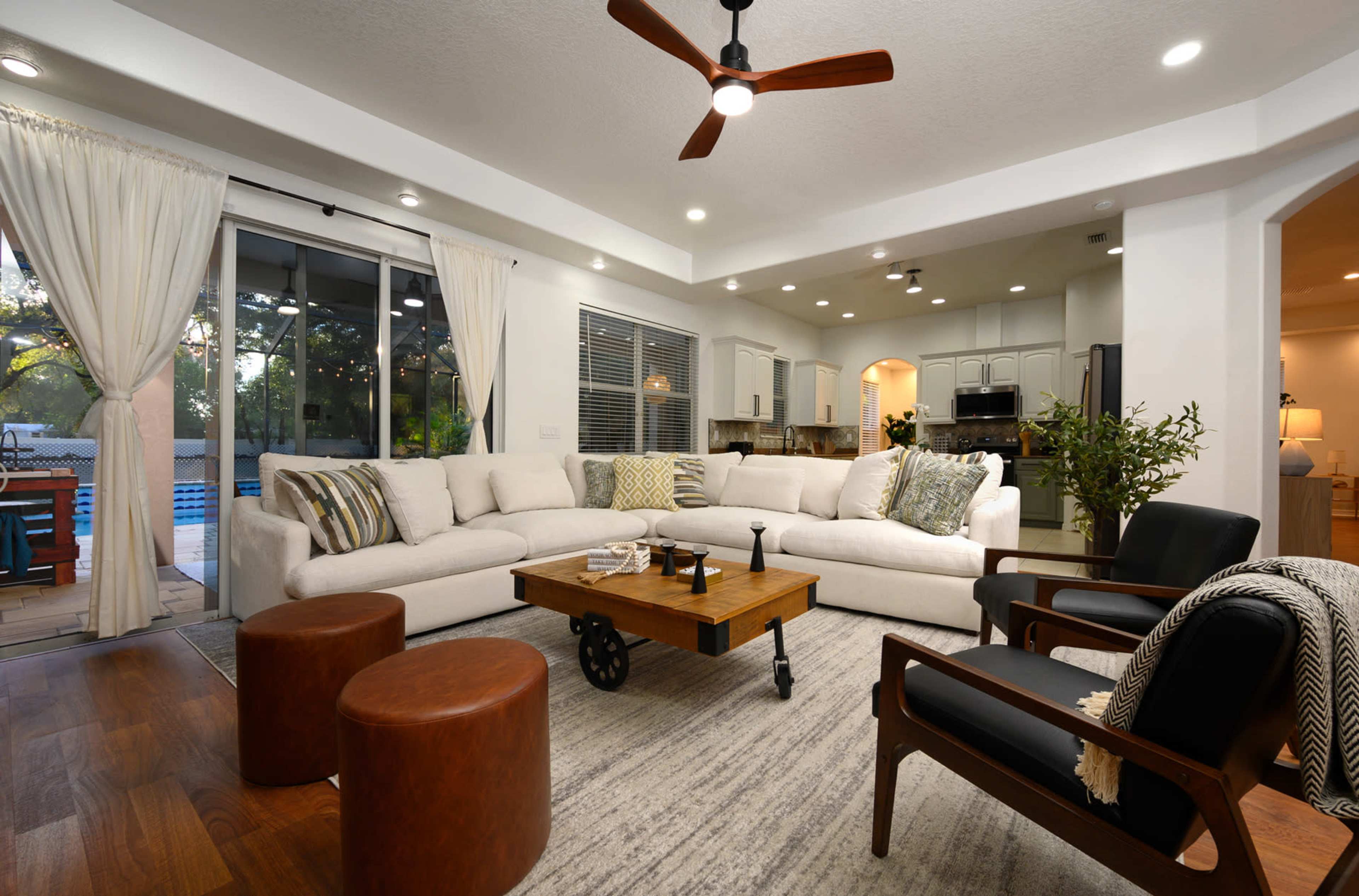 A modern living room with a large sectional sofa, two brown ottomans, and a coffee table, all arranged around a central area that leads to a kitchen visible in the background.