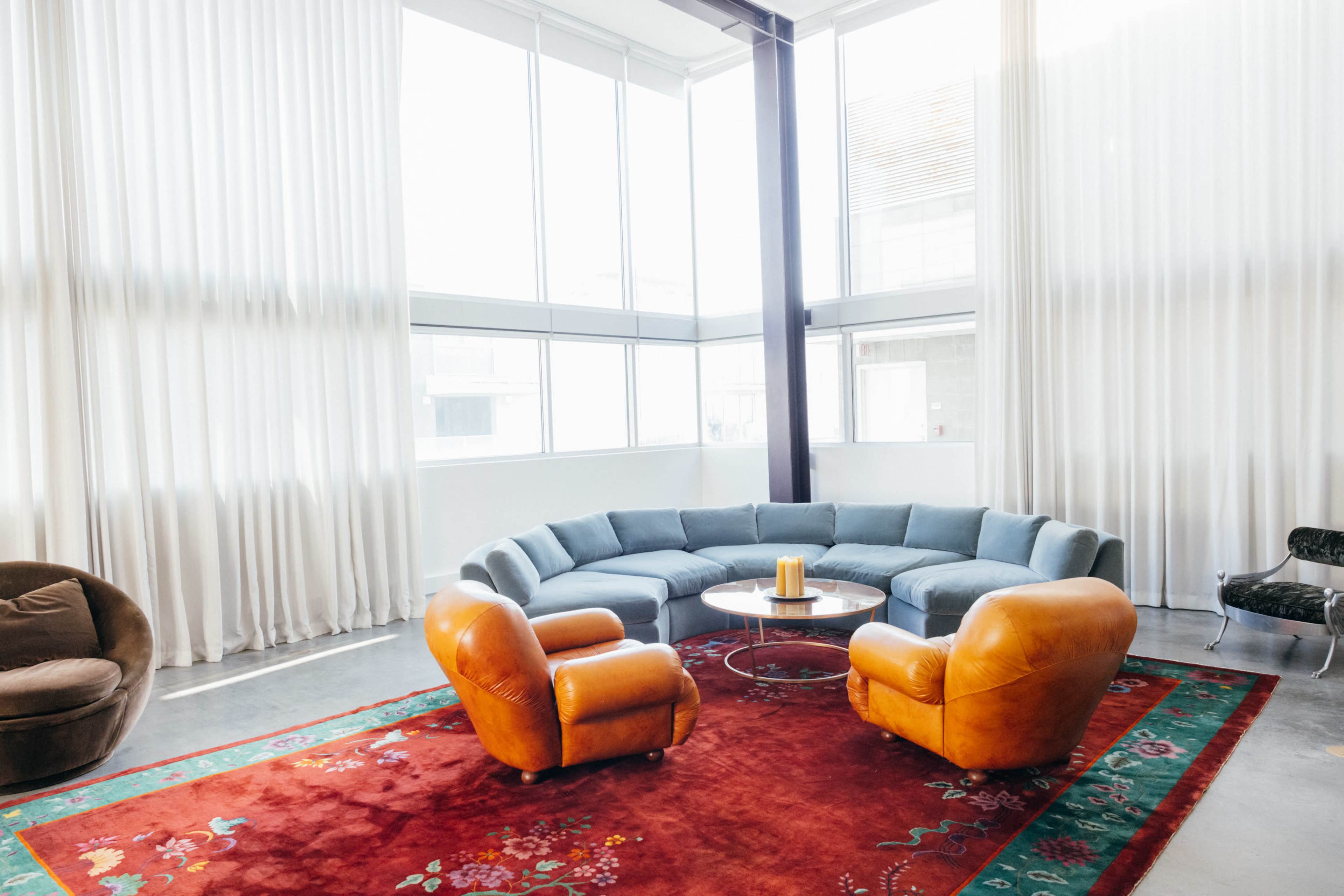 A spacious room features a curved blue sofa, two orange armchairs, a round table in the center, and large windows with sheer white curtains, all sitting atop a patterned red rug.