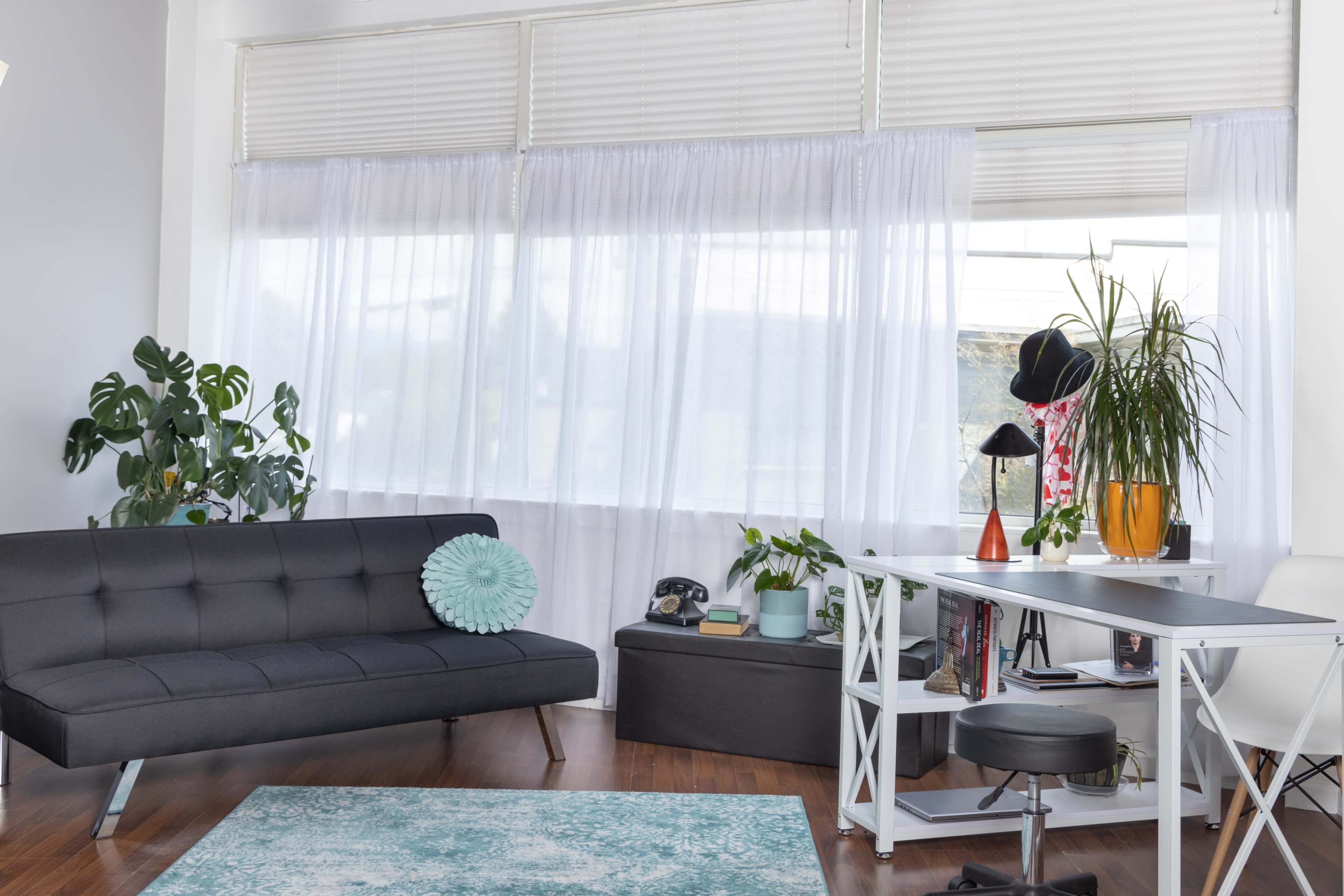 The image shows a modern workspace with a black sofa, a white desk, plants, and large windows covered with sheer curtains.