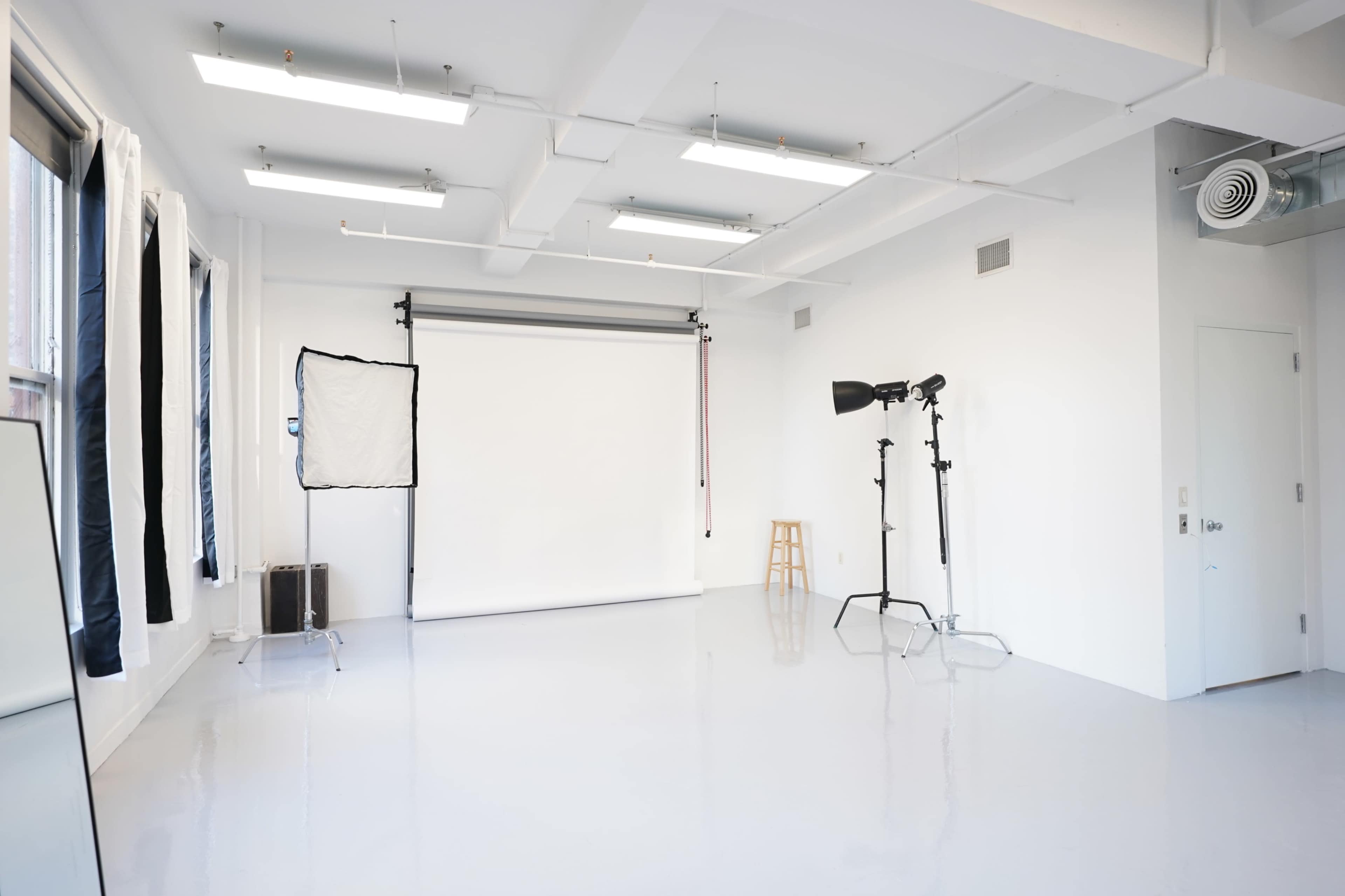 The image shows a spacious, well-lit photography studio with white walls, a backdrop setup, and lighting equipment.