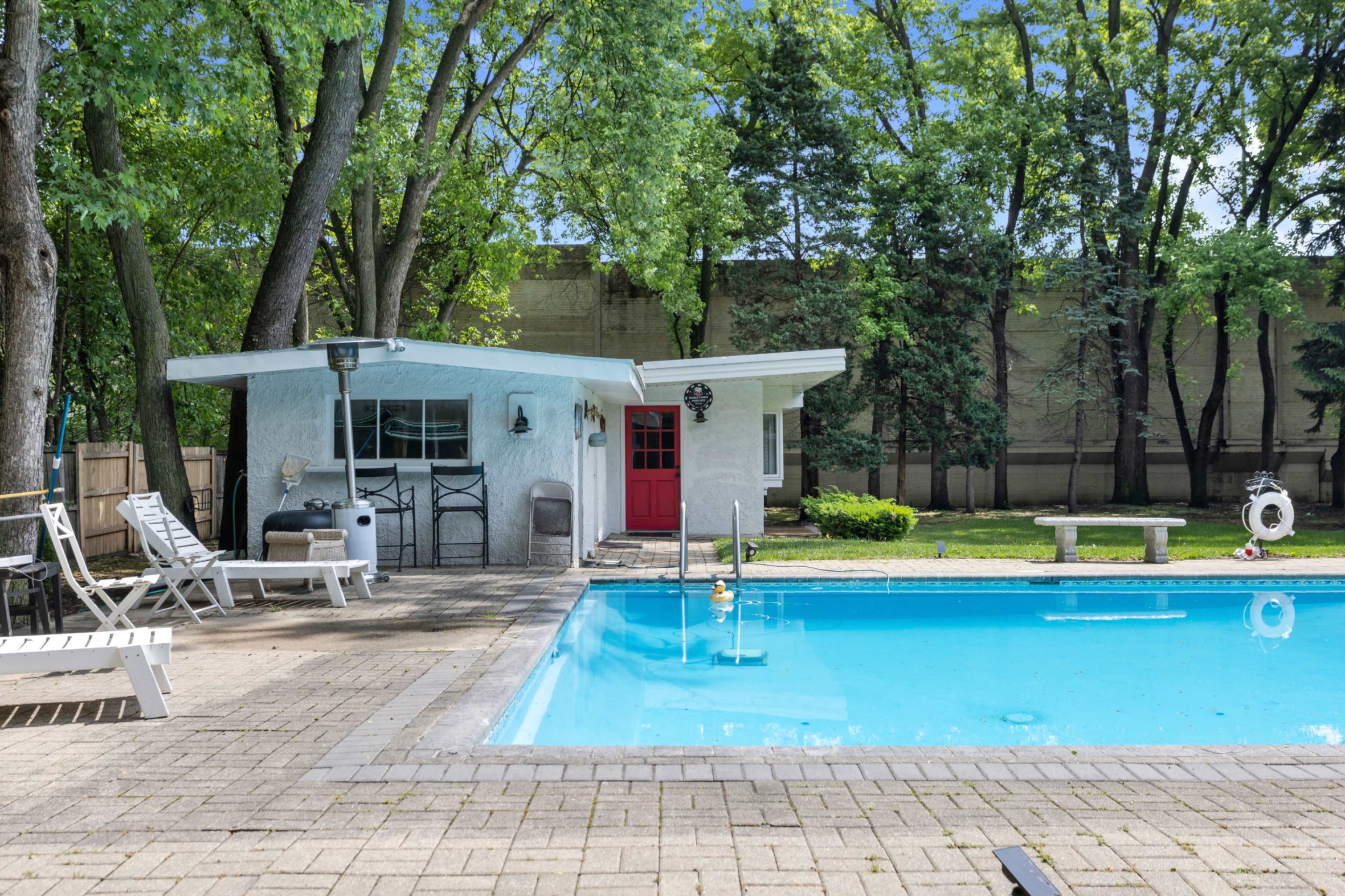 The scene features a swimming pool surrounded by lounge chairs and a small white building with a red door set among trees.