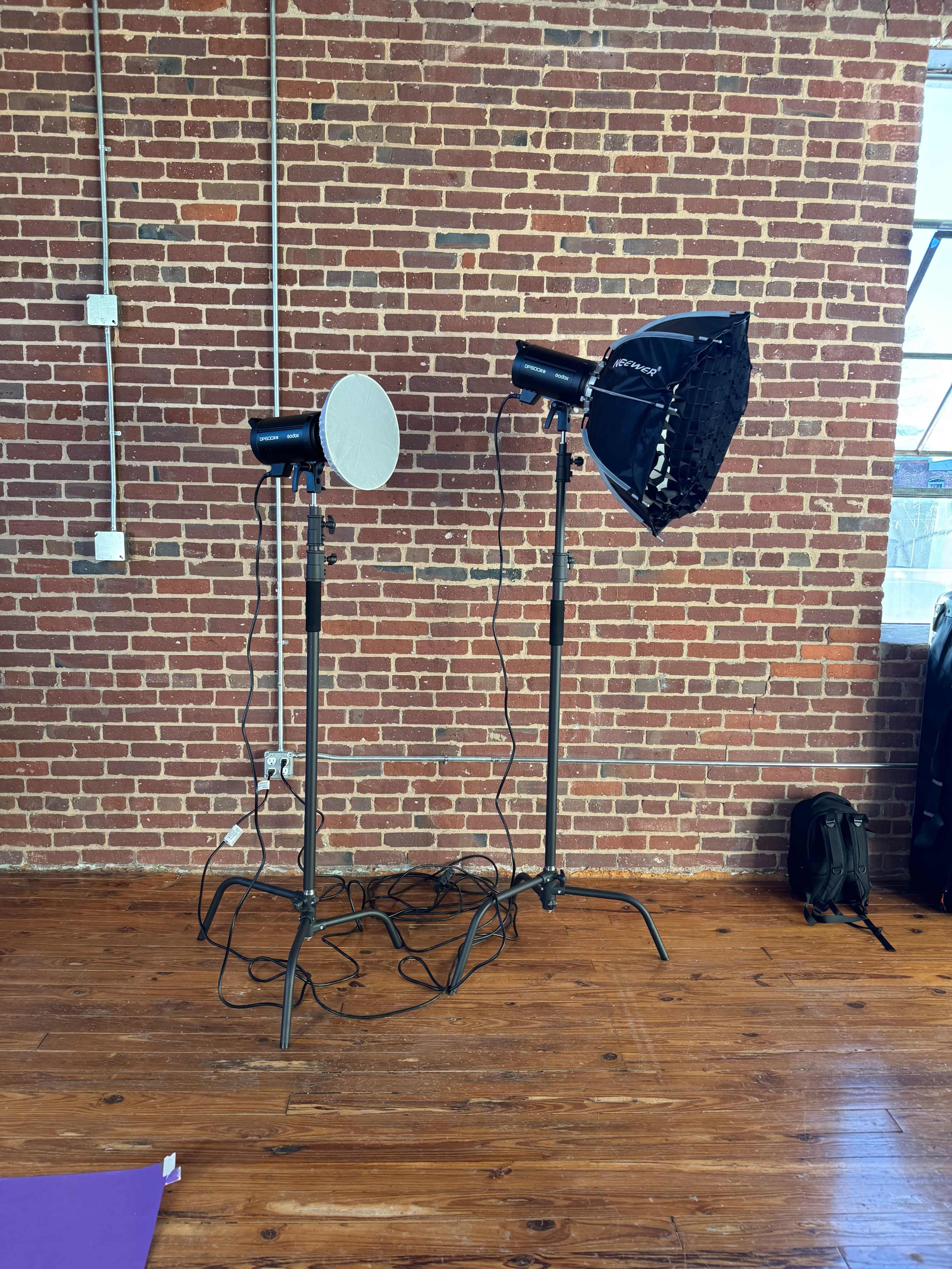 The image shows two photography lights on stands in front of a brick wall, with visible cables extending from the lights to the floor.