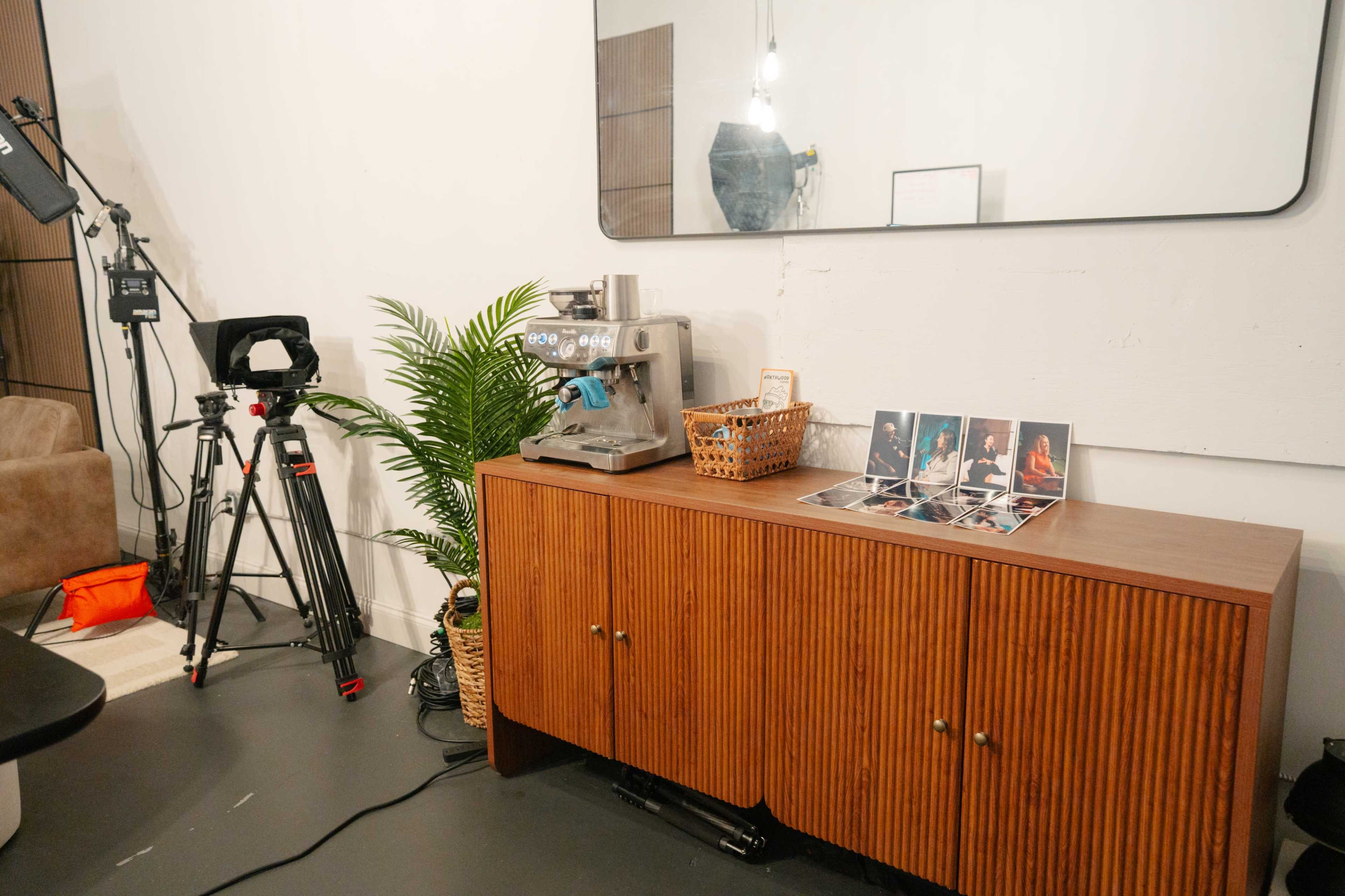 The image shows a wooden sideboard with a coffee machine and a basket on top, alongside a few photographs, in a well-lit room featuring a camera setup and a decorative plant.