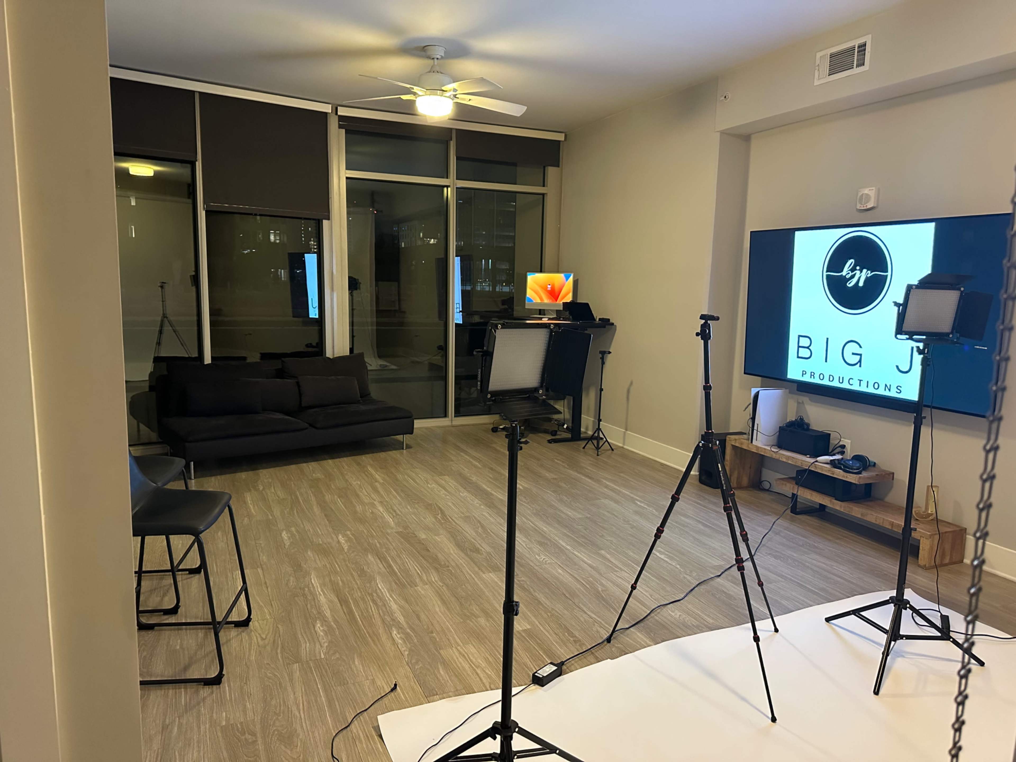 A modern studio space with a black couch, a large screen displaying "BIG J PRODUCTIONS," and several tripods with lighting equipment set up in the room.