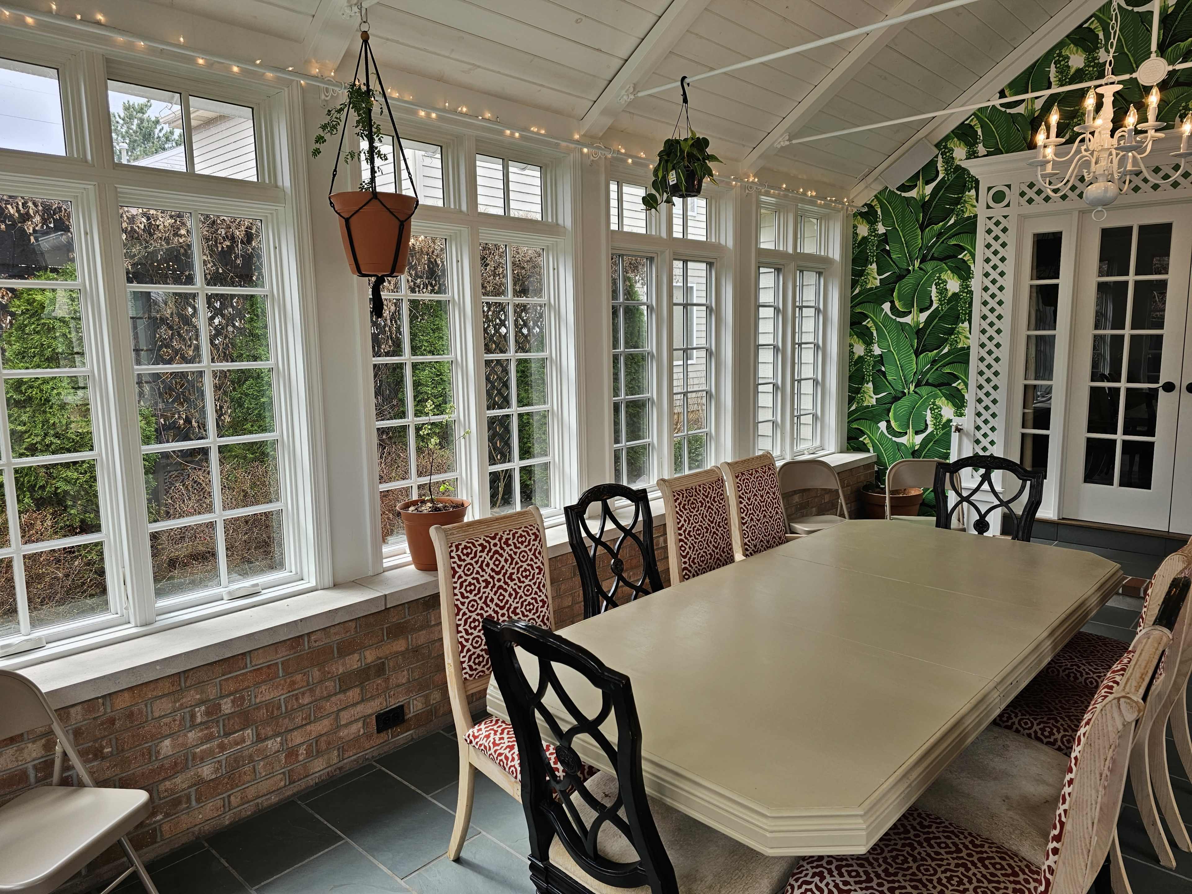 A sunlit room features a large dining table surrounded by chairs, with large windows overlooking greenery and a patterned wallpaper.