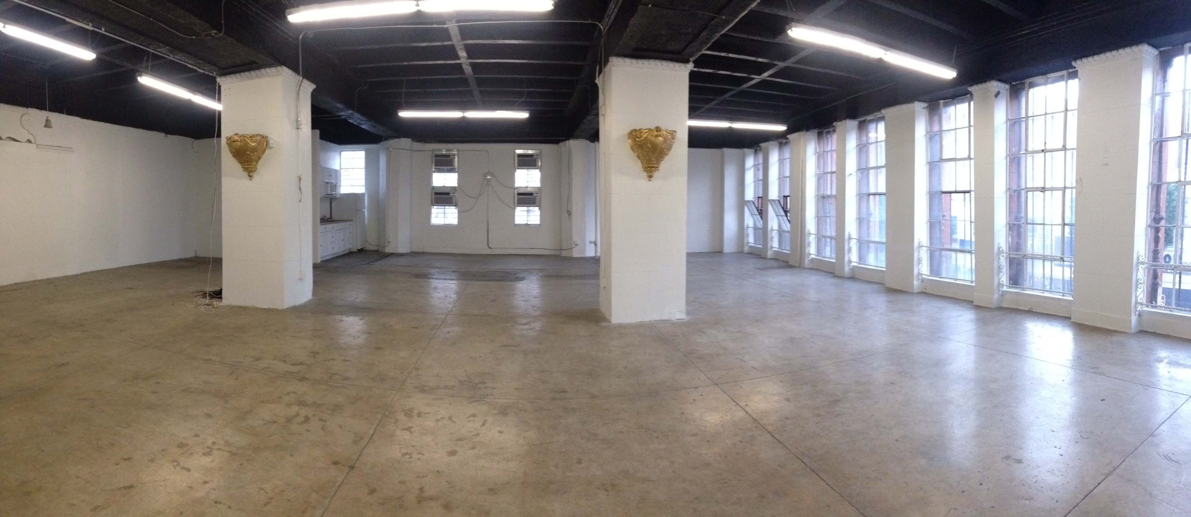 A large, empty room with concrete flooring, white walls, and large windows allowing natural light to enter.
