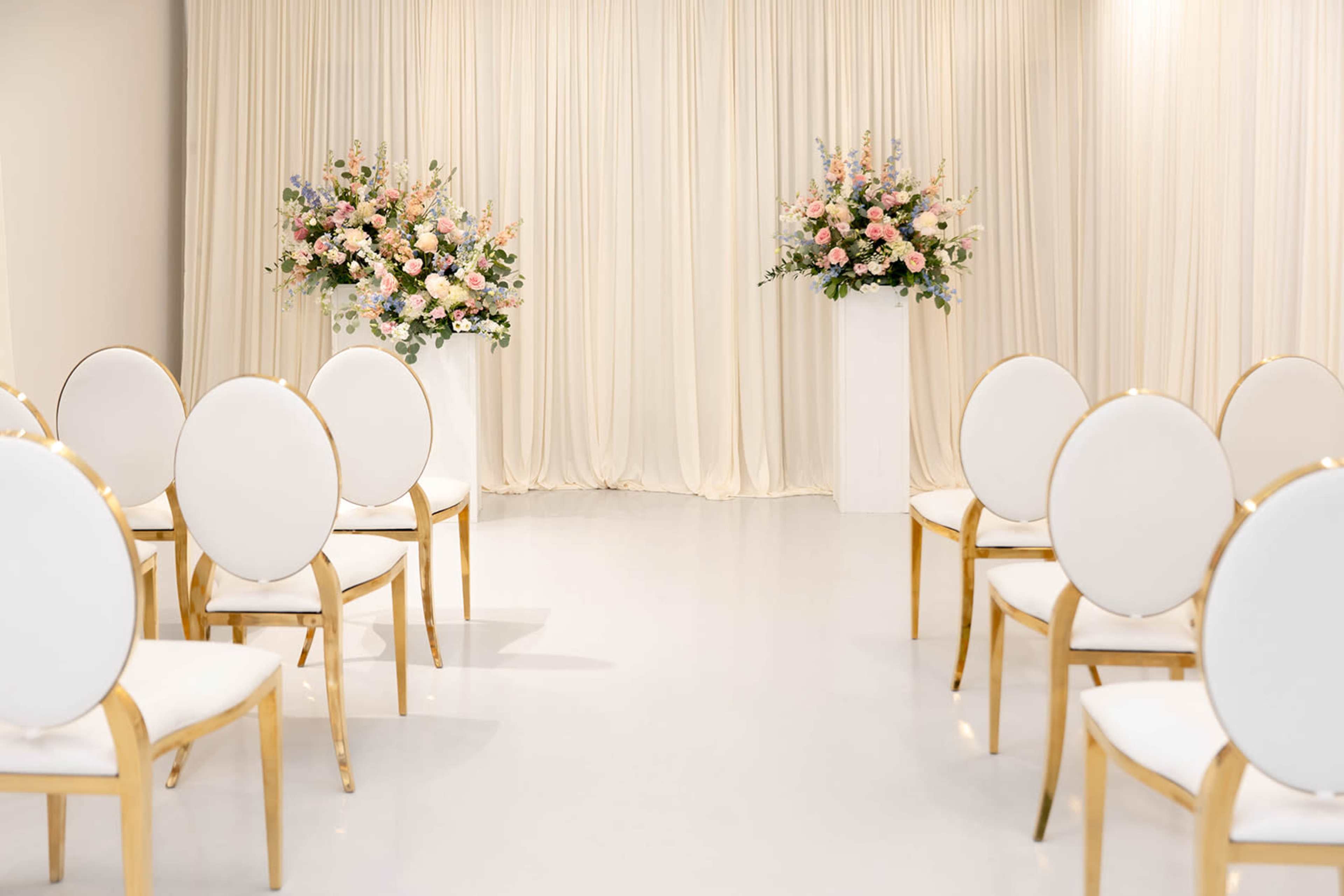 The image shows a wedding ceremony setup with white chairs arranged in rows facing a backdrop of draped fabric and floral arrangements on pedestals.