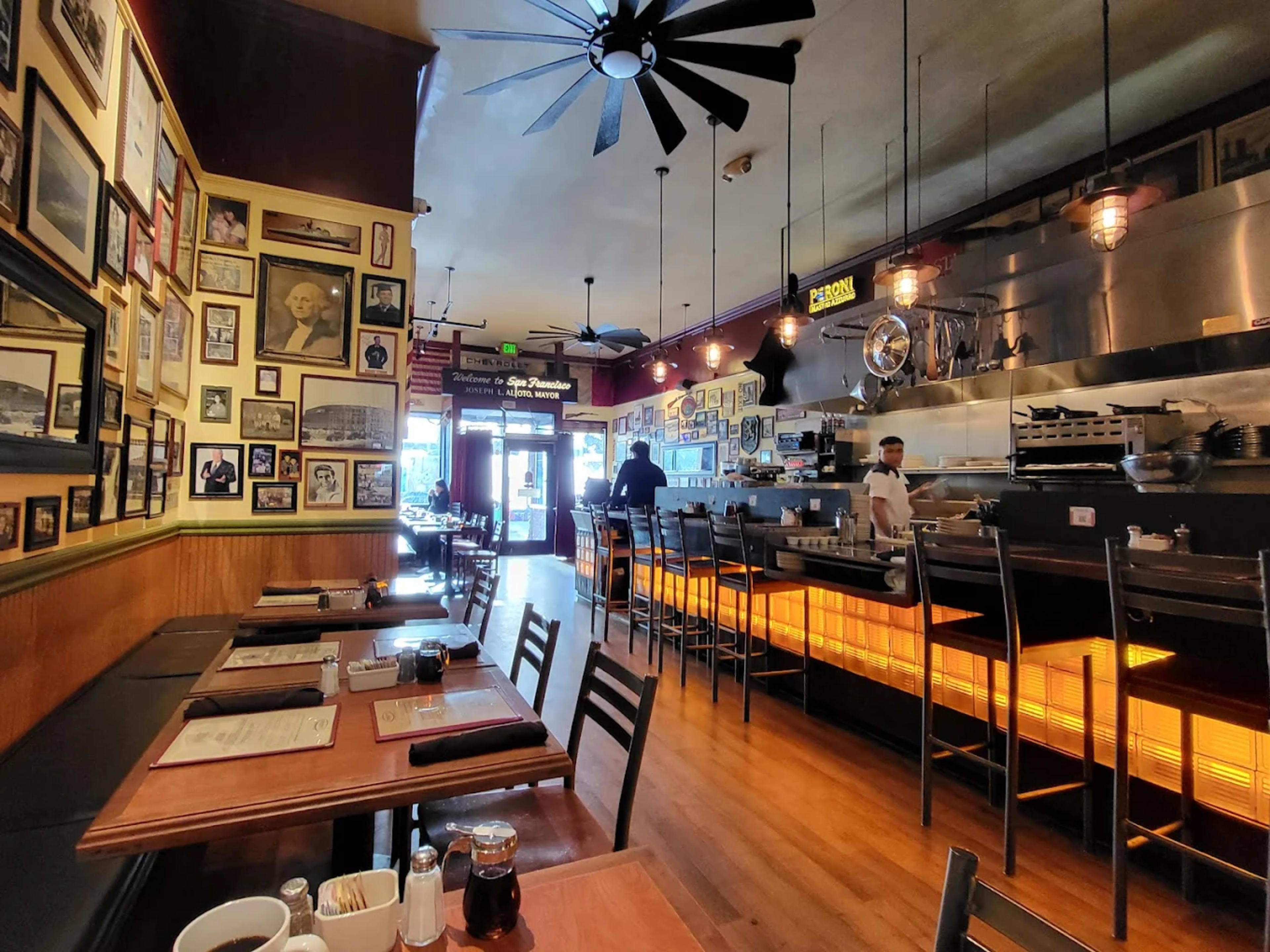 The image shows the interior of a restaurant with wooden tables, a bar area, and walls adorned with framed photographs.