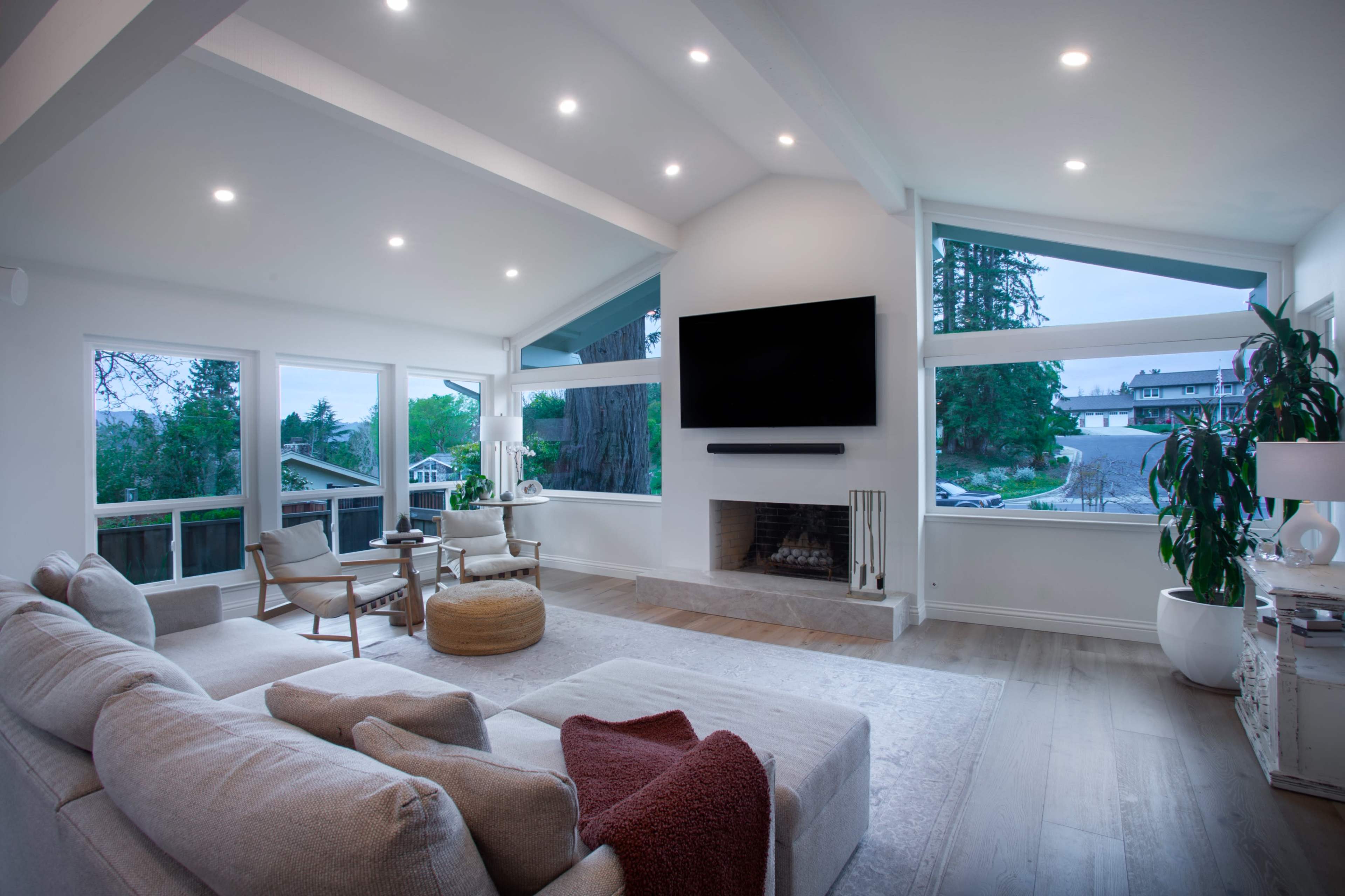 The image shows a modern living room with a light-colored sofa, two armchairs, a coffee table, and a large television mounted on the wall, surrounded by large windows offering views of the outside.