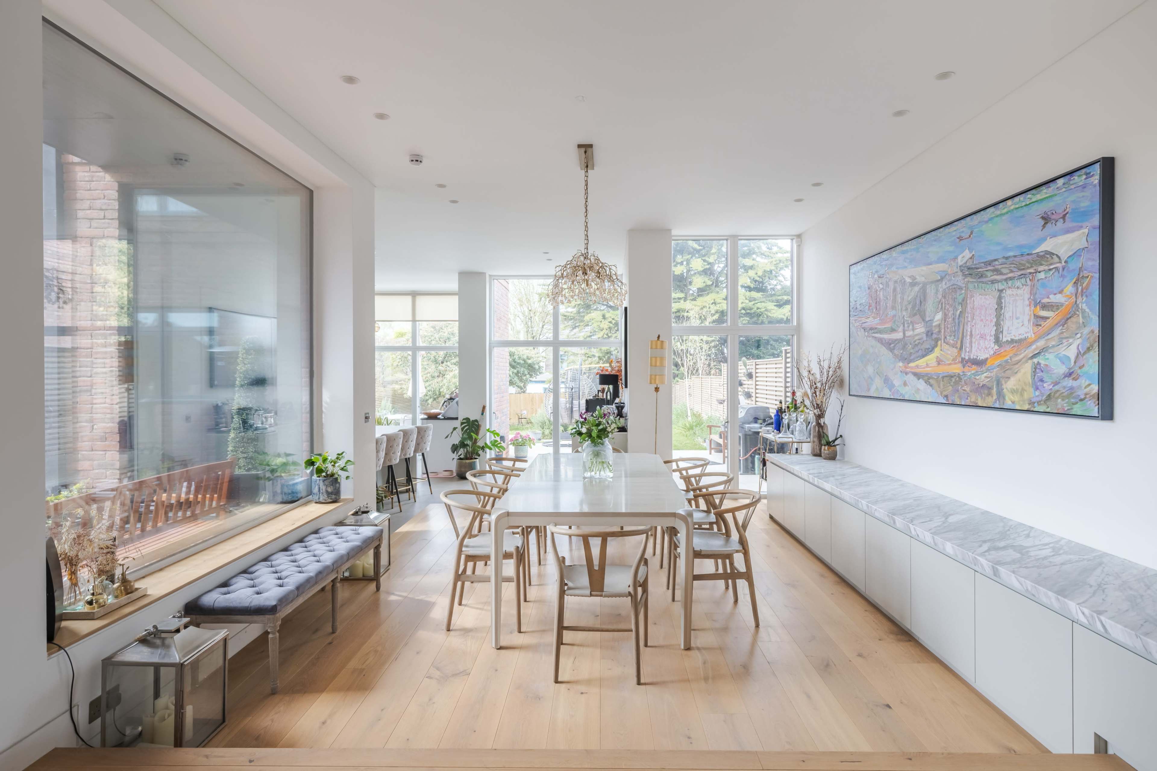 The image features a modern dining area with a central table surrounded by chairs, large windows, and a vibrant piece of artwork on the wall.