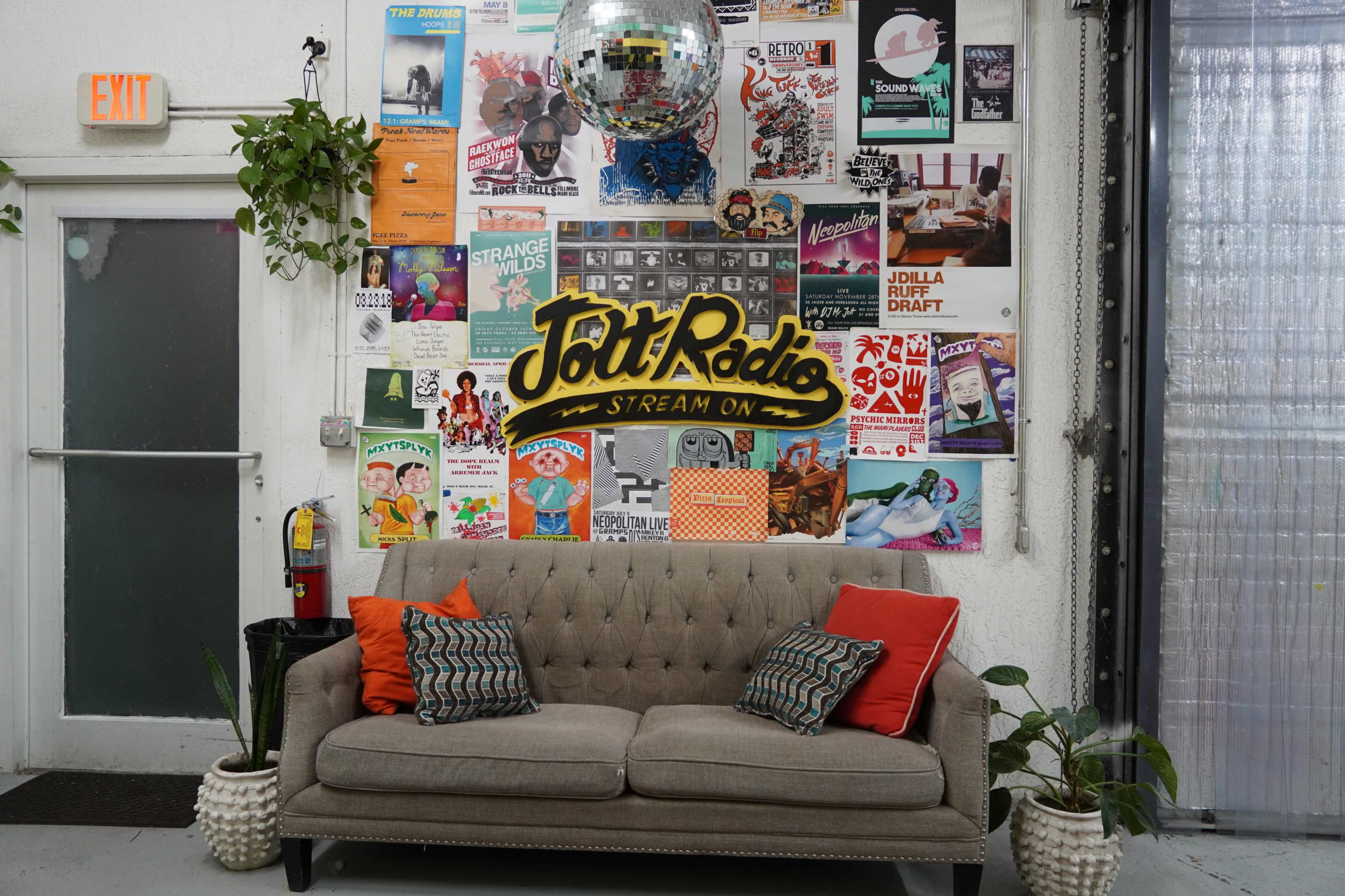 A cozy seating area with a beige couch adorned with orange pillows, set against a collage of colorful posters on a wall, featuring the phrase "Jolt Radio" prominently displayed.