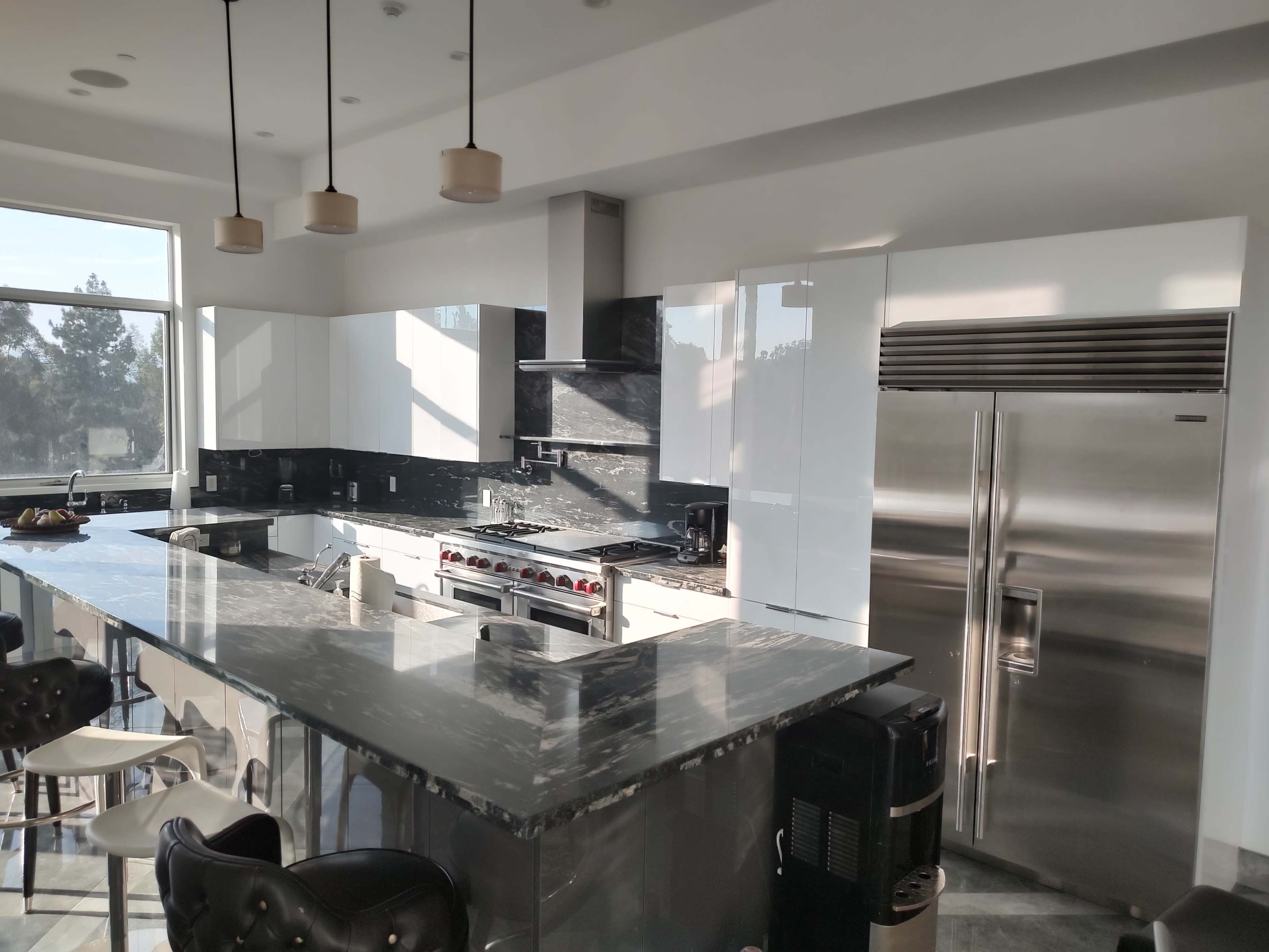 A modern kitchen with sleek white cabinets, a large marble countertop, stainless steel appliances, and a view through a large window.