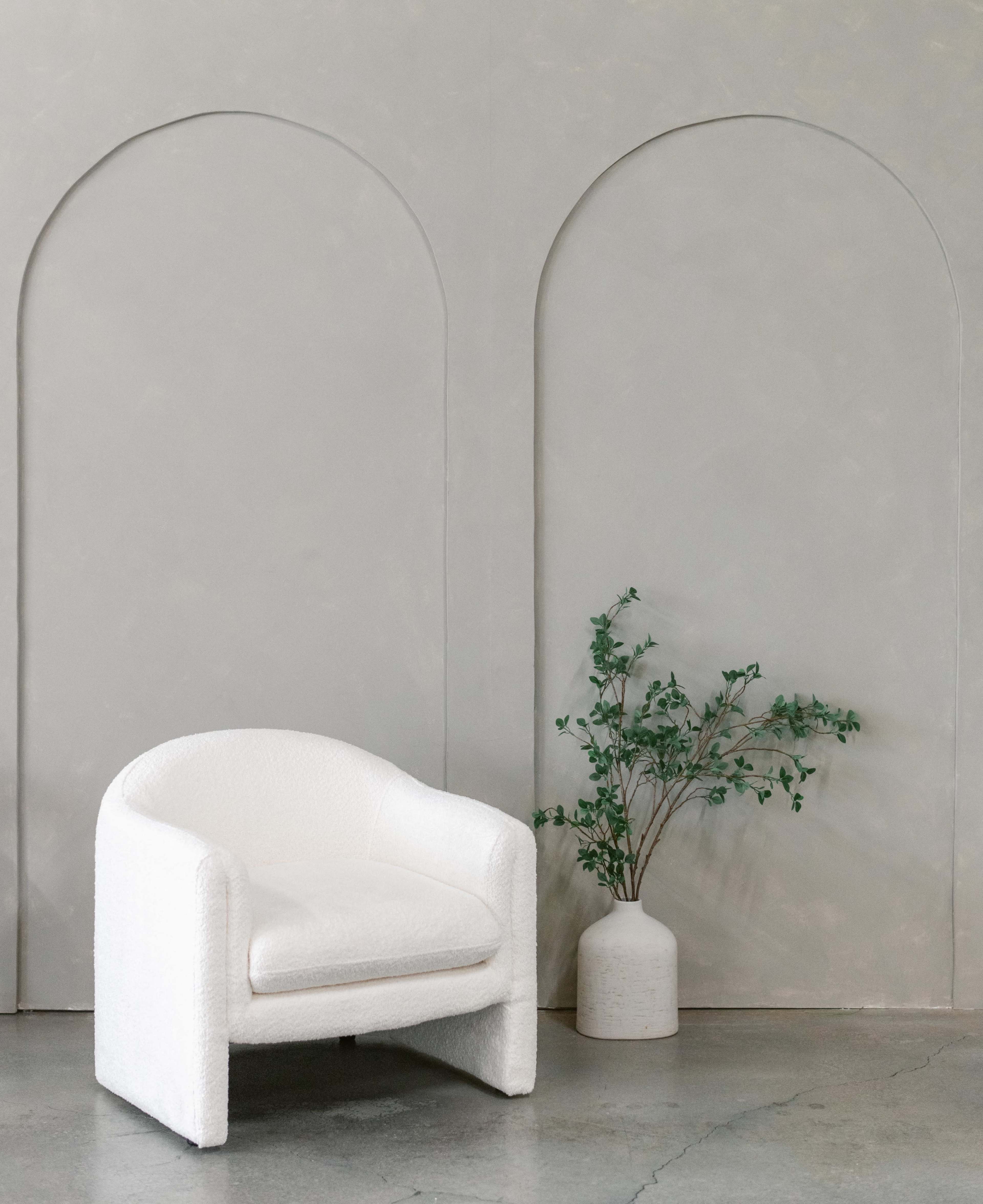 The scene features a white upholstered armchair next to a white vase with greenery against a textured gray wall with arched cutouts.