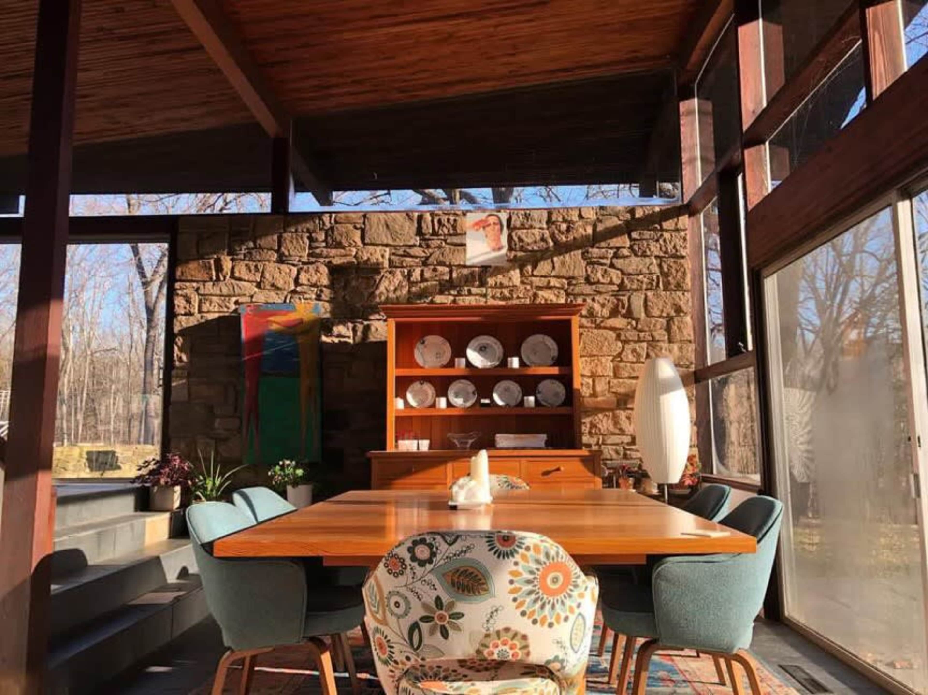A dining area features a wooden table surrounded by upholstered chairs, with a stone wall and large windows that allow natural light to flood the space.