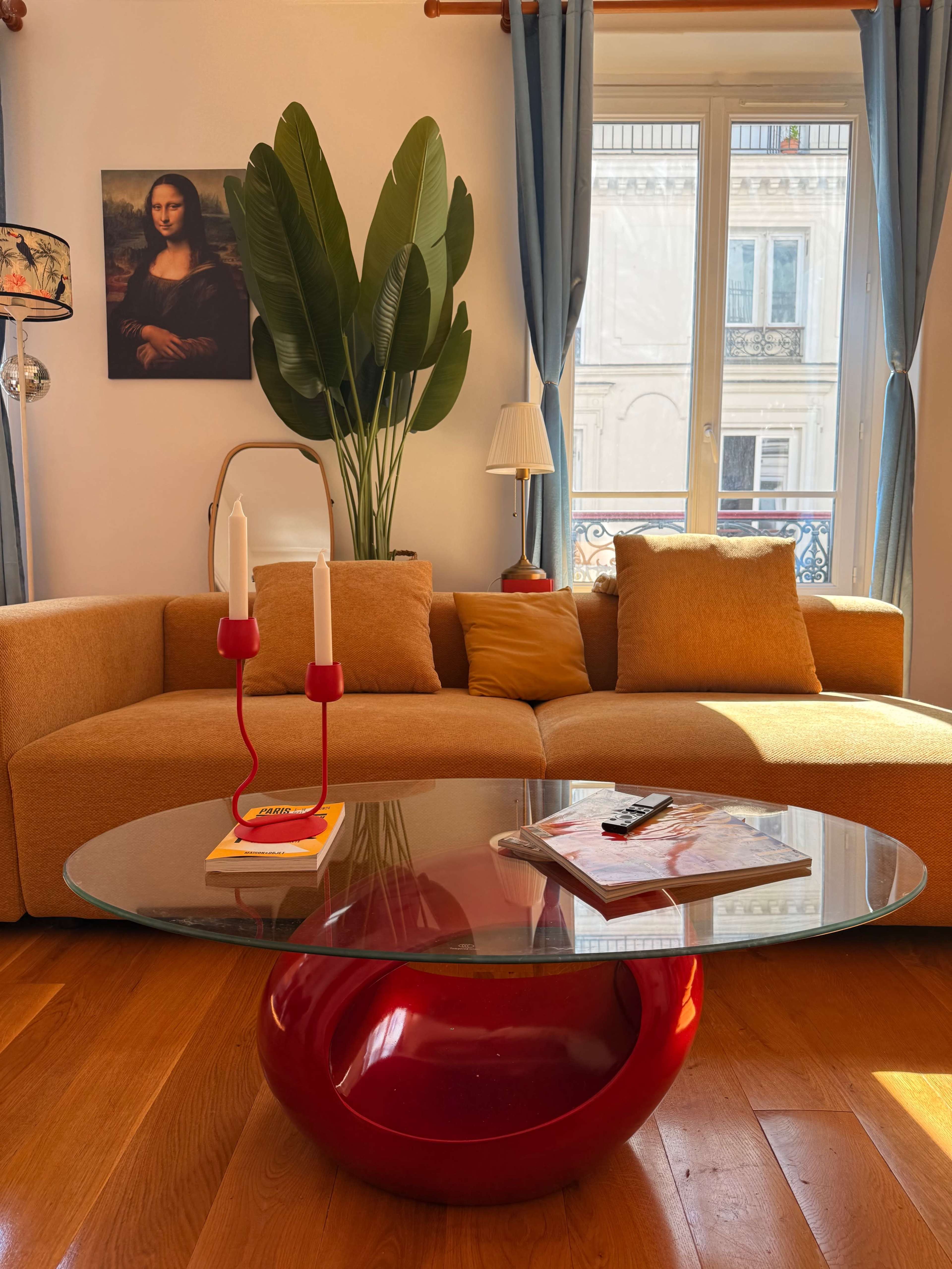 A modern living room features a beige sofa, a glass coffee table with a red base, and a large plant beside a window, with the Mona Lisa portrait hanging on the wall.