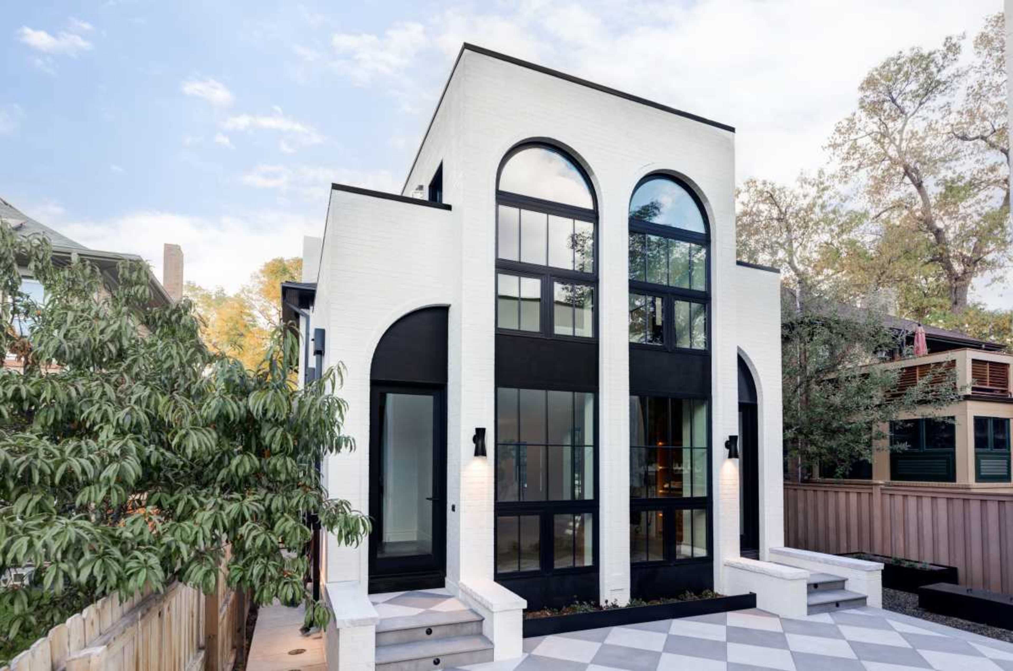 The image shows a modern two-story house with a white brick facade, large black-framed windows, and arched doorways.