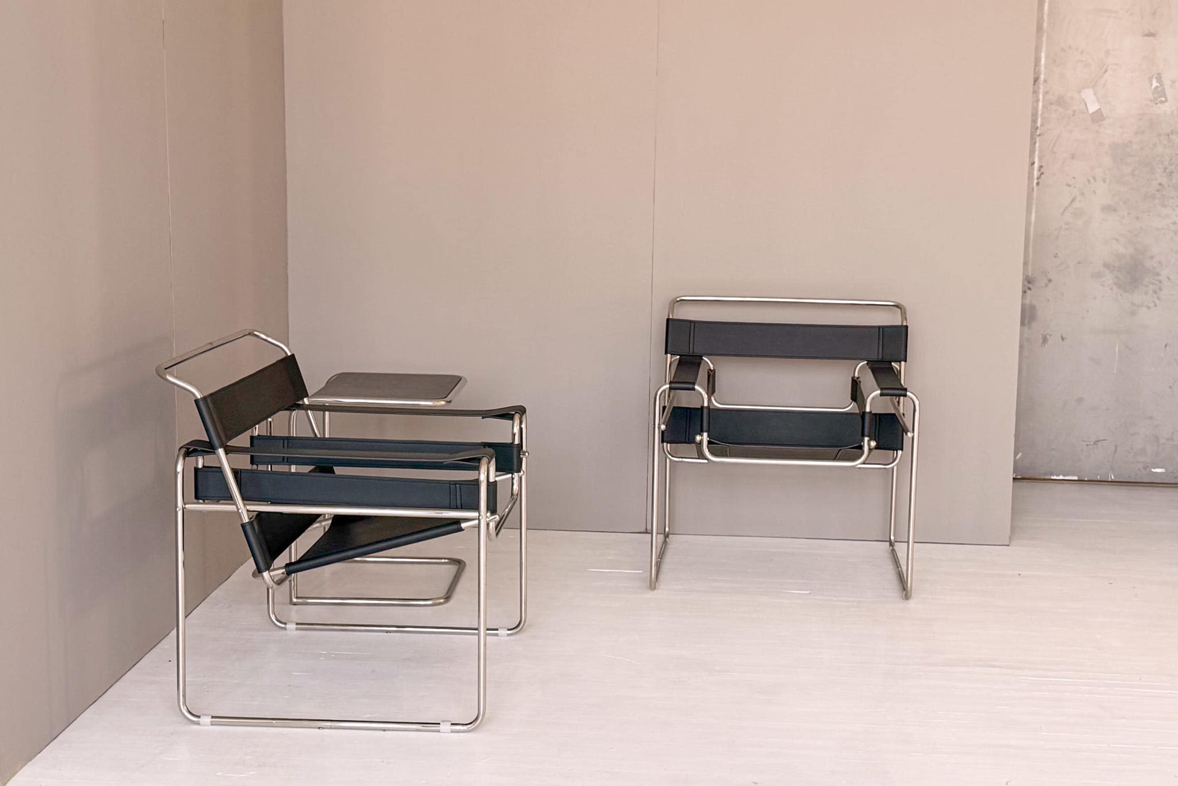 Two minimalist metal chairs with black fabric seats are positioned next to a small table against a plain, light-colored wall.