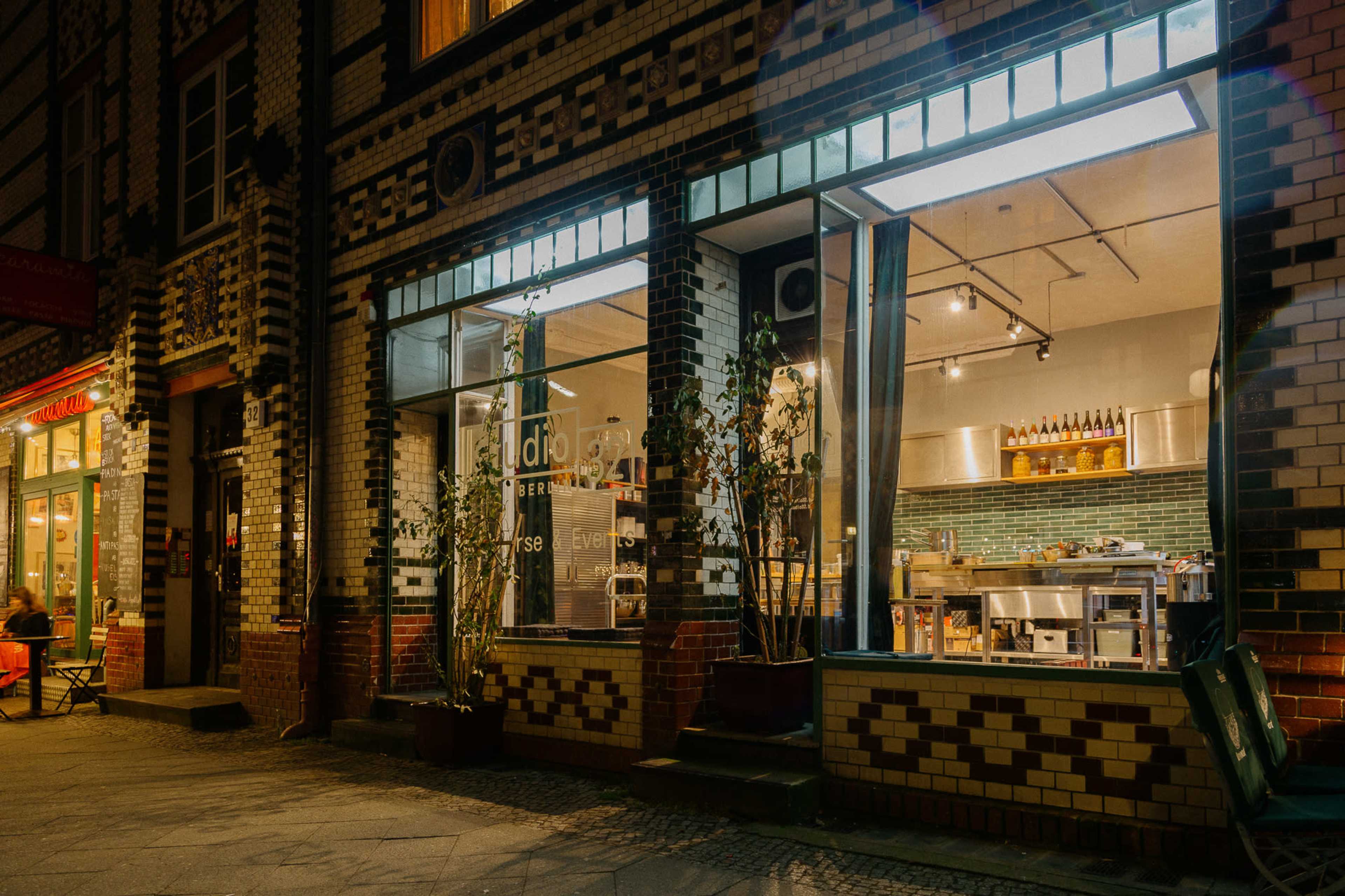 A restaurant with large front windows displays its interior and kitchen, featuring plants in pots at the entrance, against a backdrop of patterned brickwork.