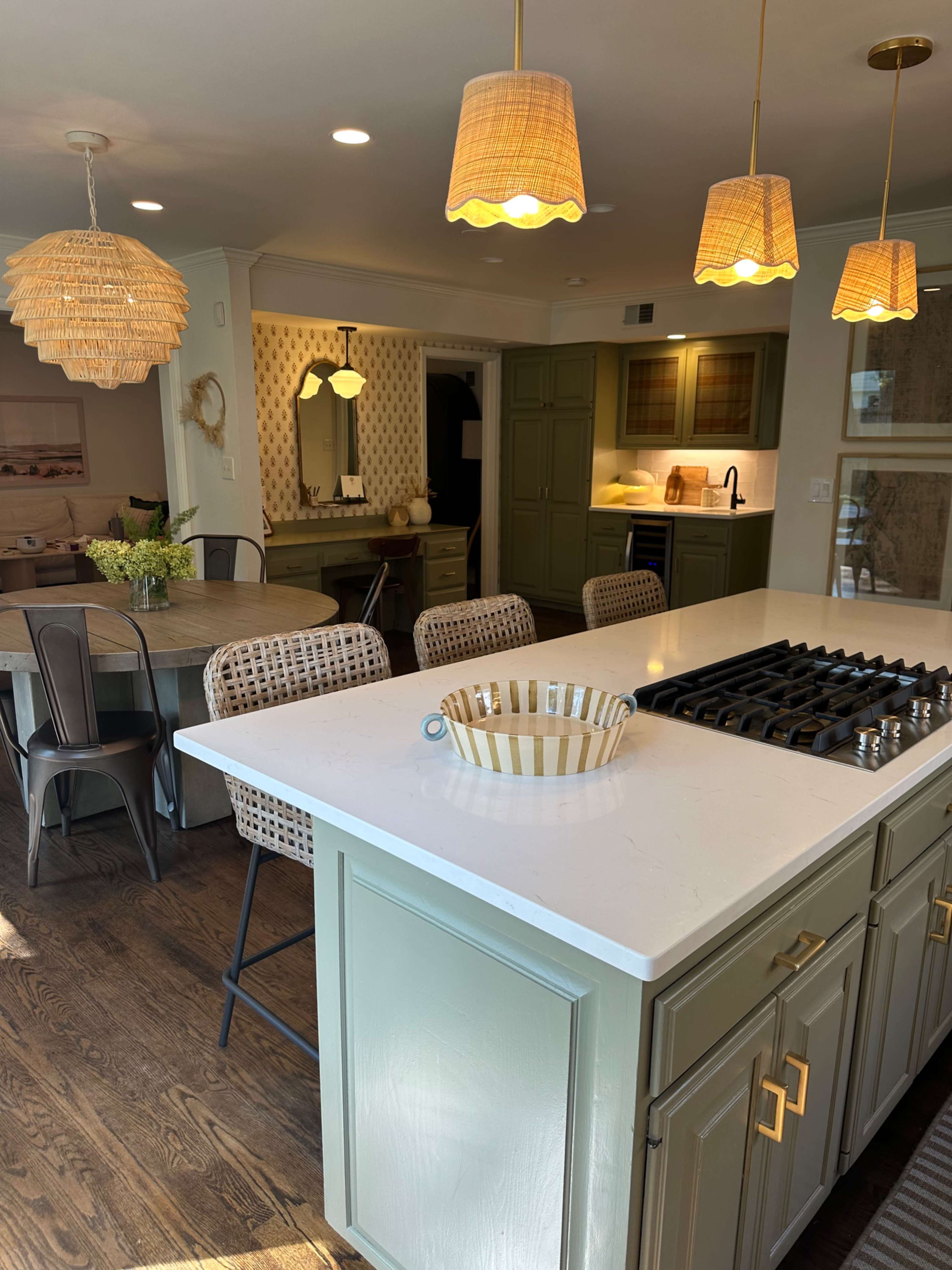 The image shows a modern kitchen featuring a white countertop, green cabinetry, a gas stove, and pendant lighting, with a dining area visible in the background.