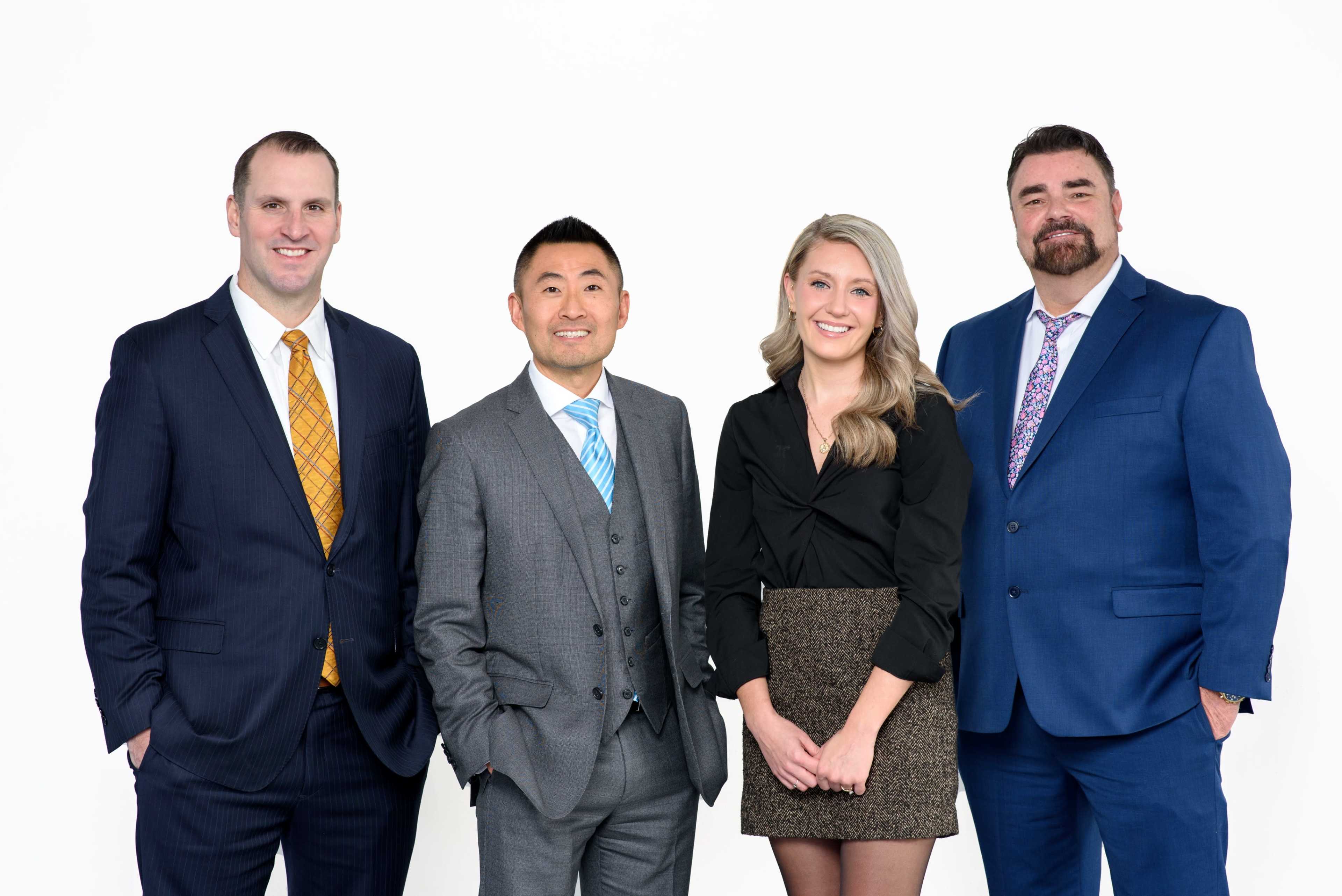 A group of four professionals stands in front of a plain white background, dressed in business attire.