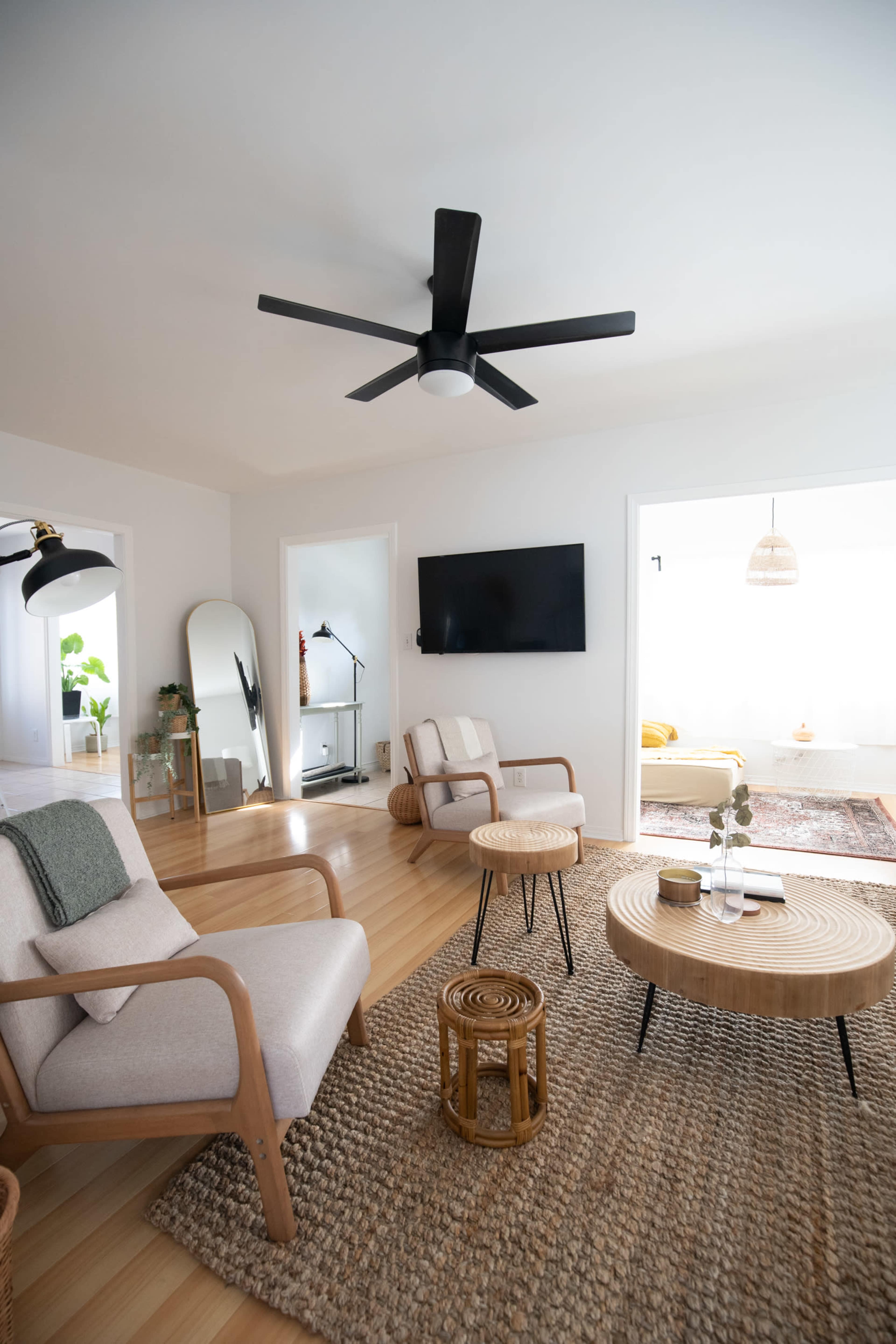 The living room features two wooden-framed chairs, a round coffee table, and a textured rug, with a ceiling fan above and a television mounted on the wall.