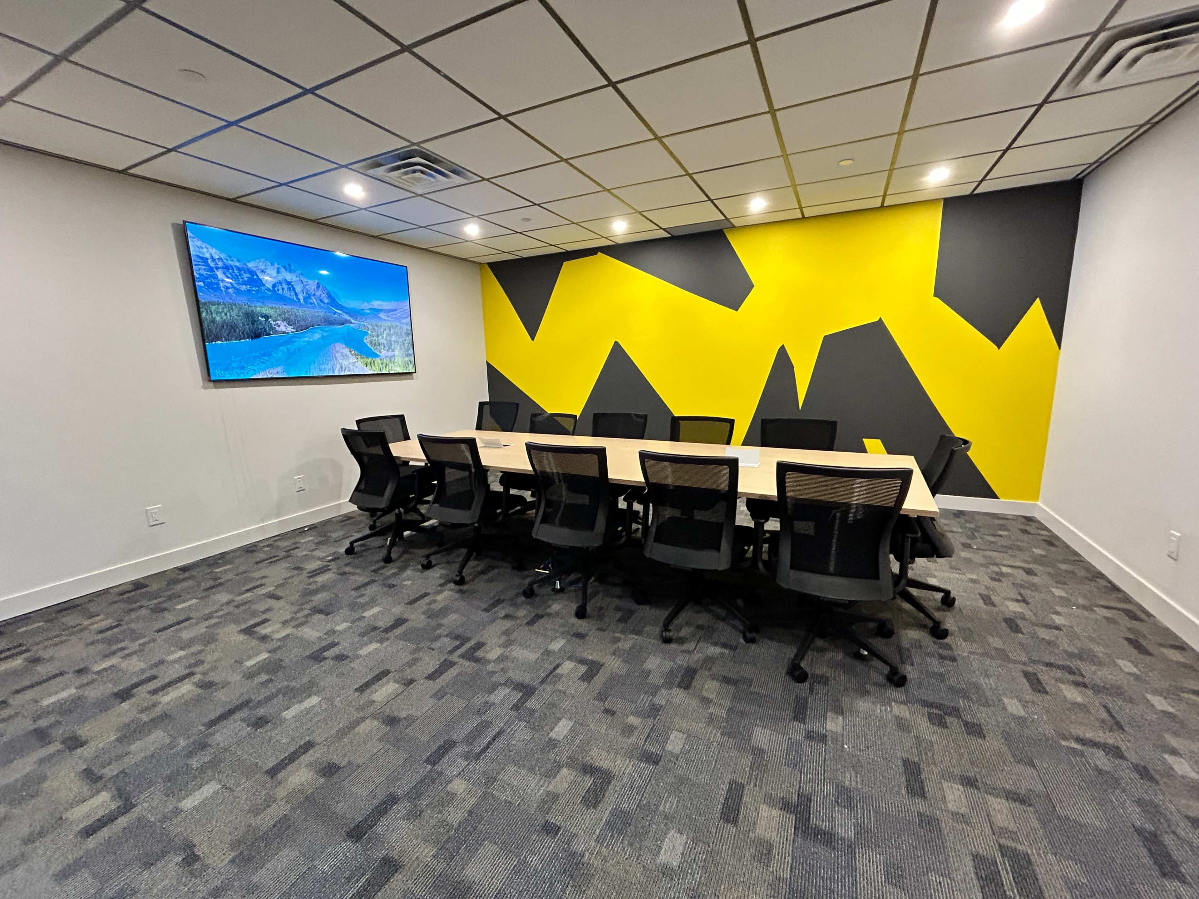 A modern conference room features a long table surrounded by black chairs, with a large screen on one wall and a vibrant yellow and gray geometric mural.