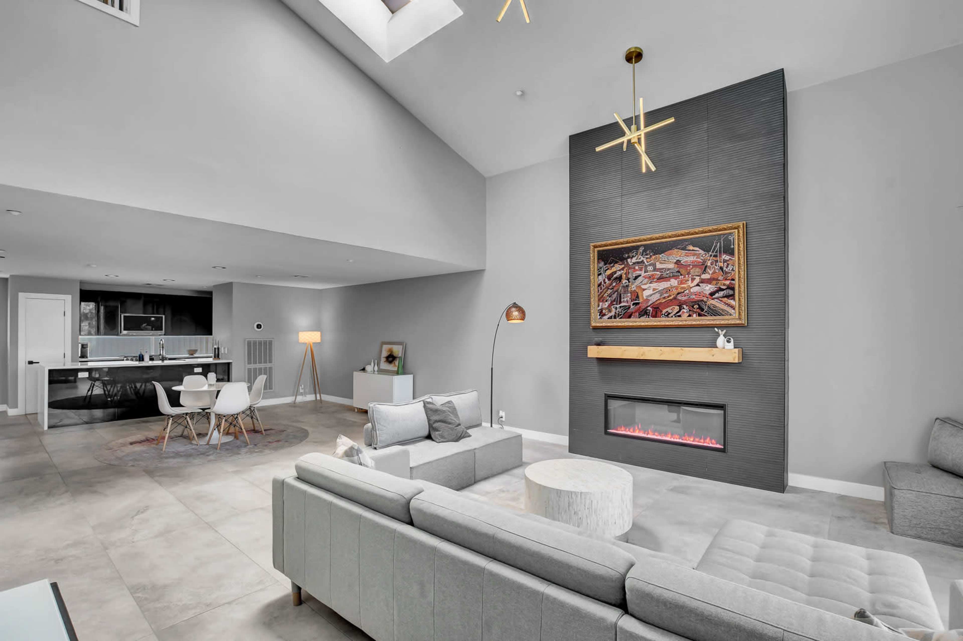 The image shows a modern living room featuring a gray sectional sofa, a round coffee table, and a large wall-mounted painting above a sleek electric fireplace.