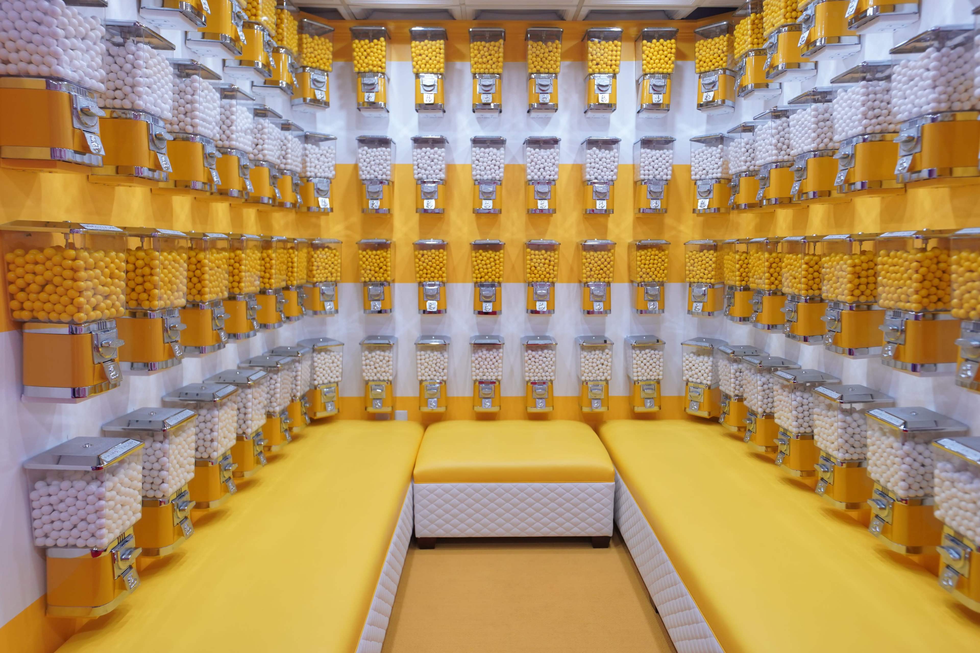 A brightly colored room filled with multiple dispensers containing yellow and white spherical candies arranged systematically along the walls.