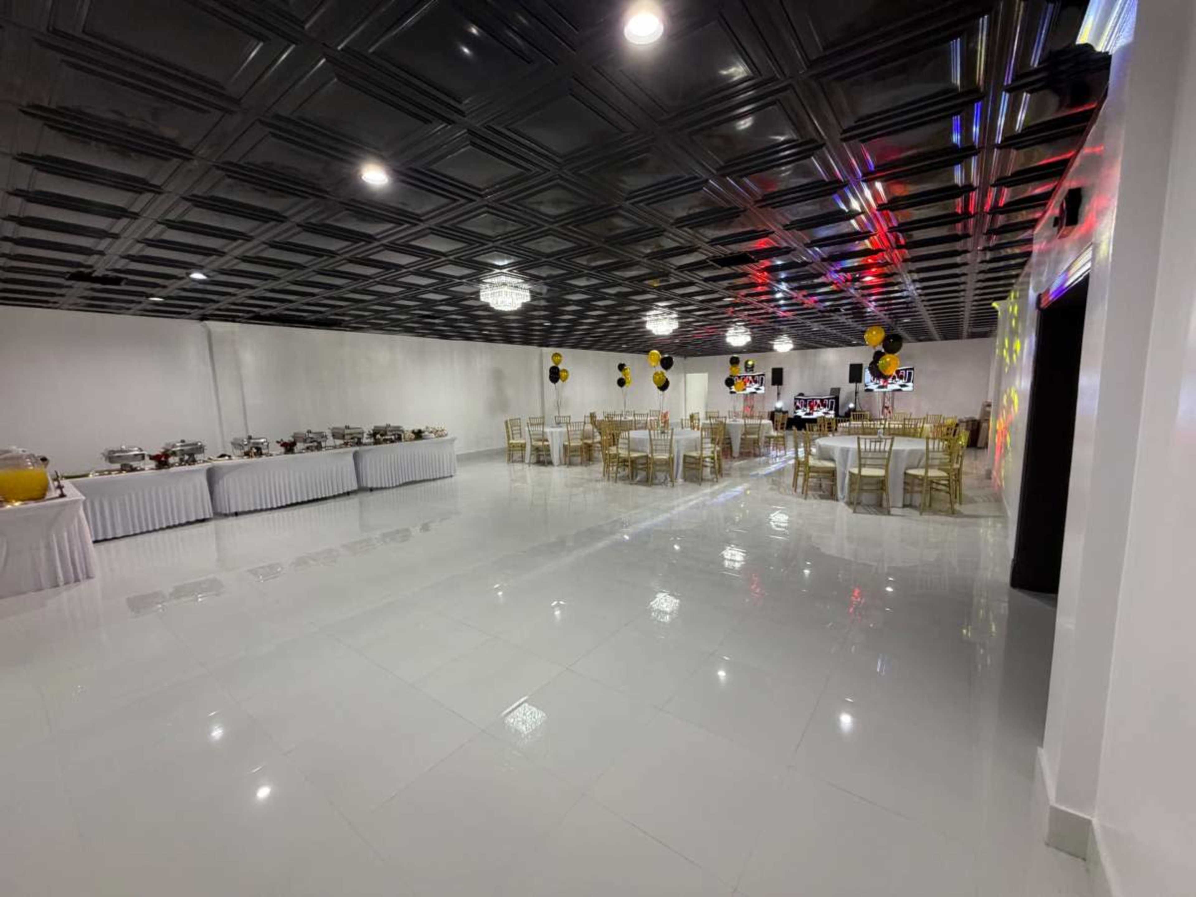 The image shows a spacious event hall with polished white floors, decorated tables, and a buffet setup along one side, featuring yellow and black balloons.