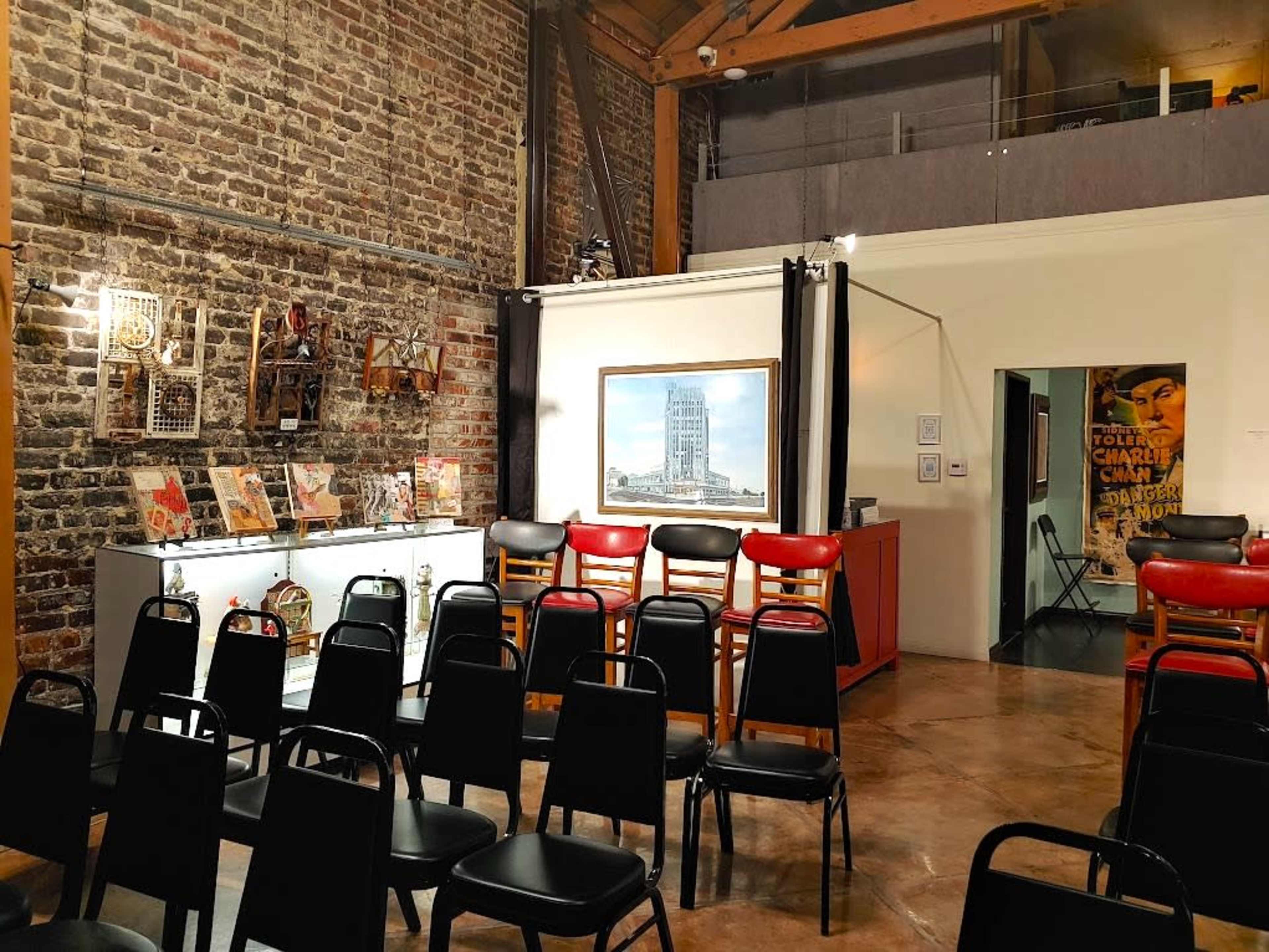 The image shows an interior space with exposed brick walls, rows of black chairs facing a display case, and a large framed artwork on the wall.