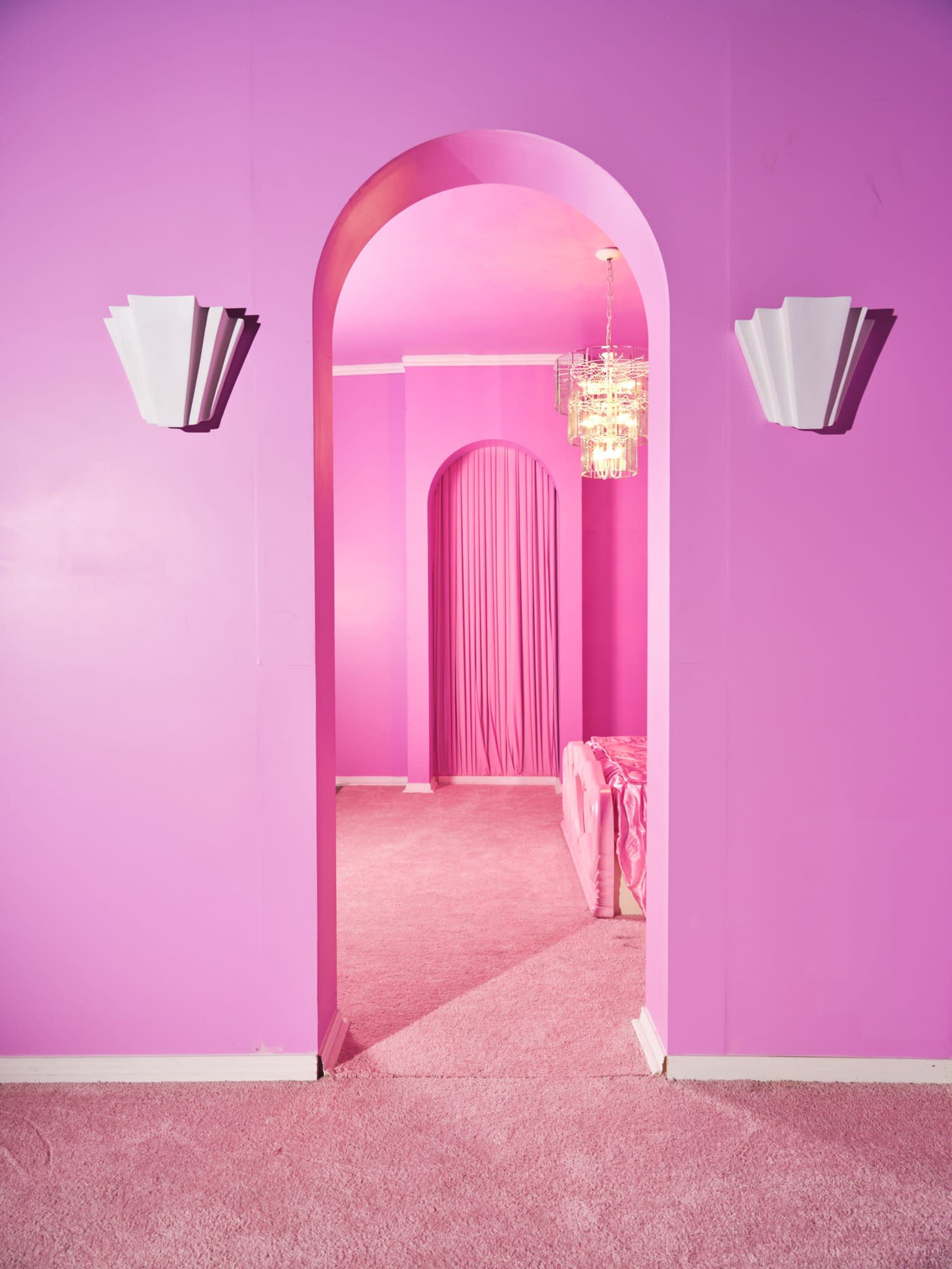 A hallway with pink walls and a carpet features two archways, a chandelier, and wall sconces.