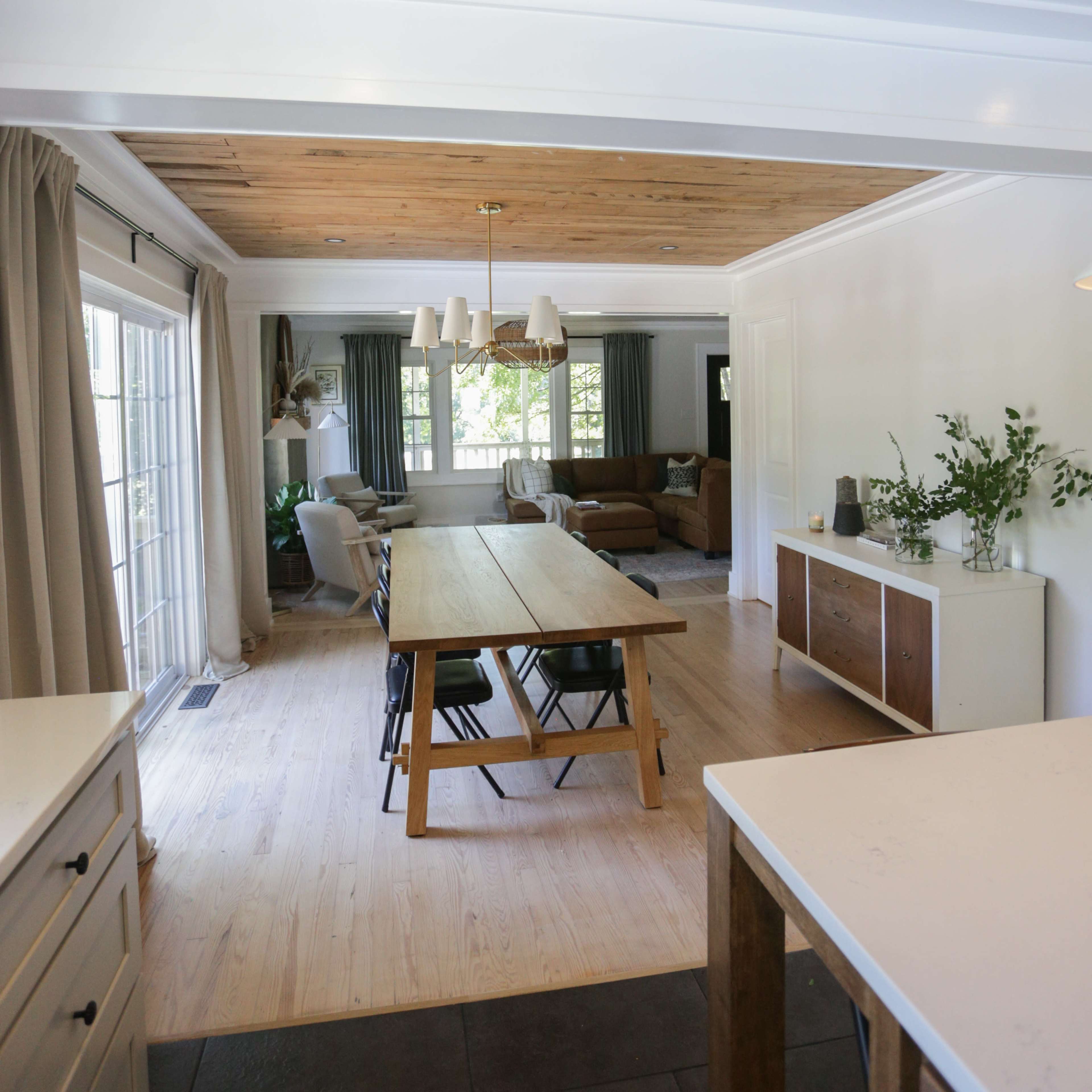 The image shows a bright, open-concept dining area leading into a living room, featuring a wooden table and chairs, a sideboard, and large windows allowing natural light in.