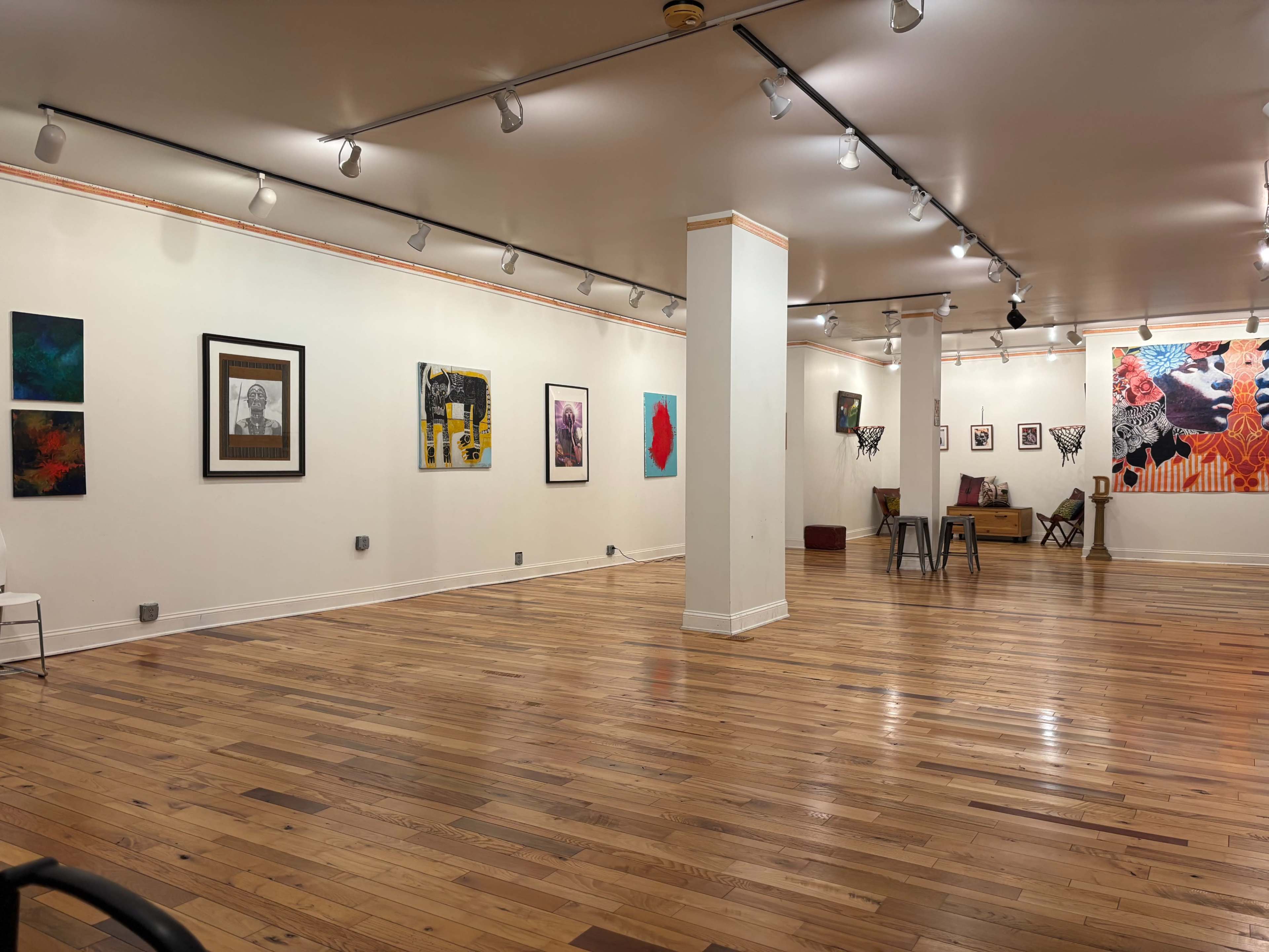 The image shows a spacious art gallery with wooden flooring, featuring various framed artworks on the white walls and some seating arrangements in the background.