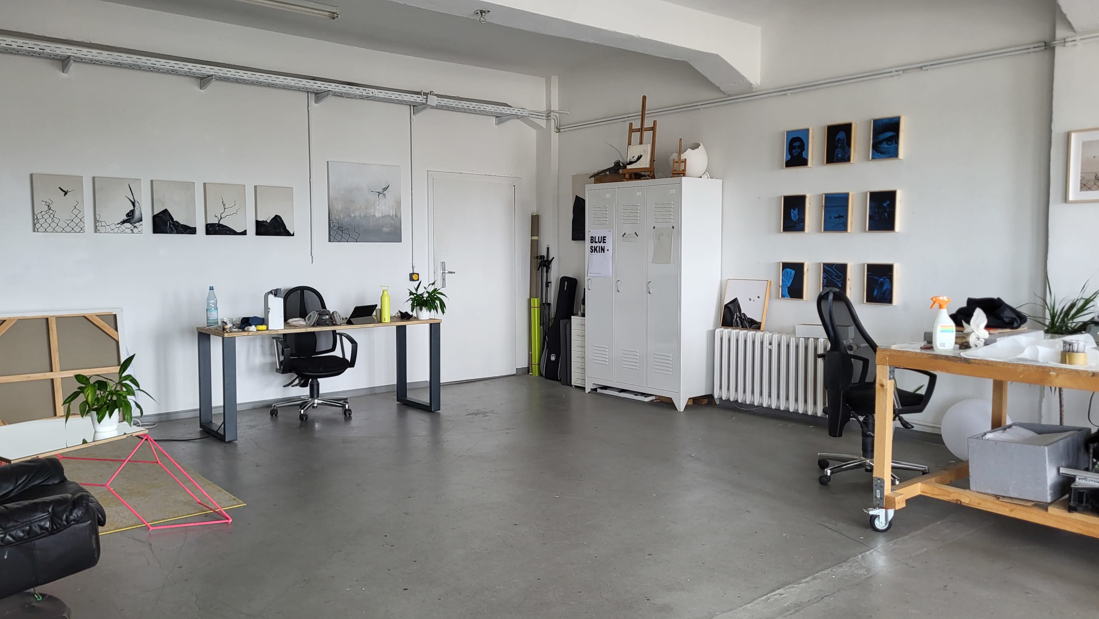 The image displays a minimalist office space featuring two desks with chairs, a white storage cabinet, framed artwork on the walls, and various plants placed in the room.