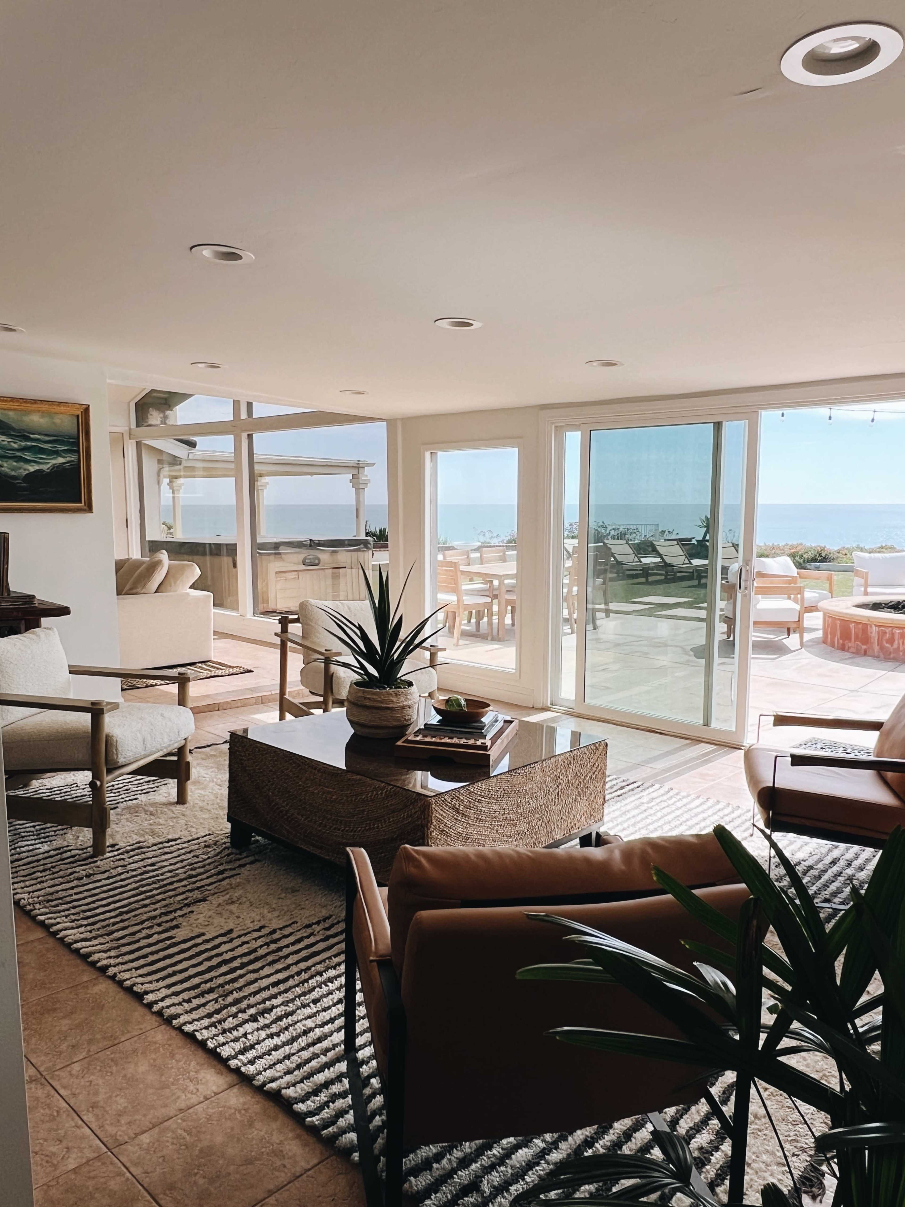 A spacious living room features comfortable seating, a central coffee table, and large windows overlooking an outdoor patio and ocean view.