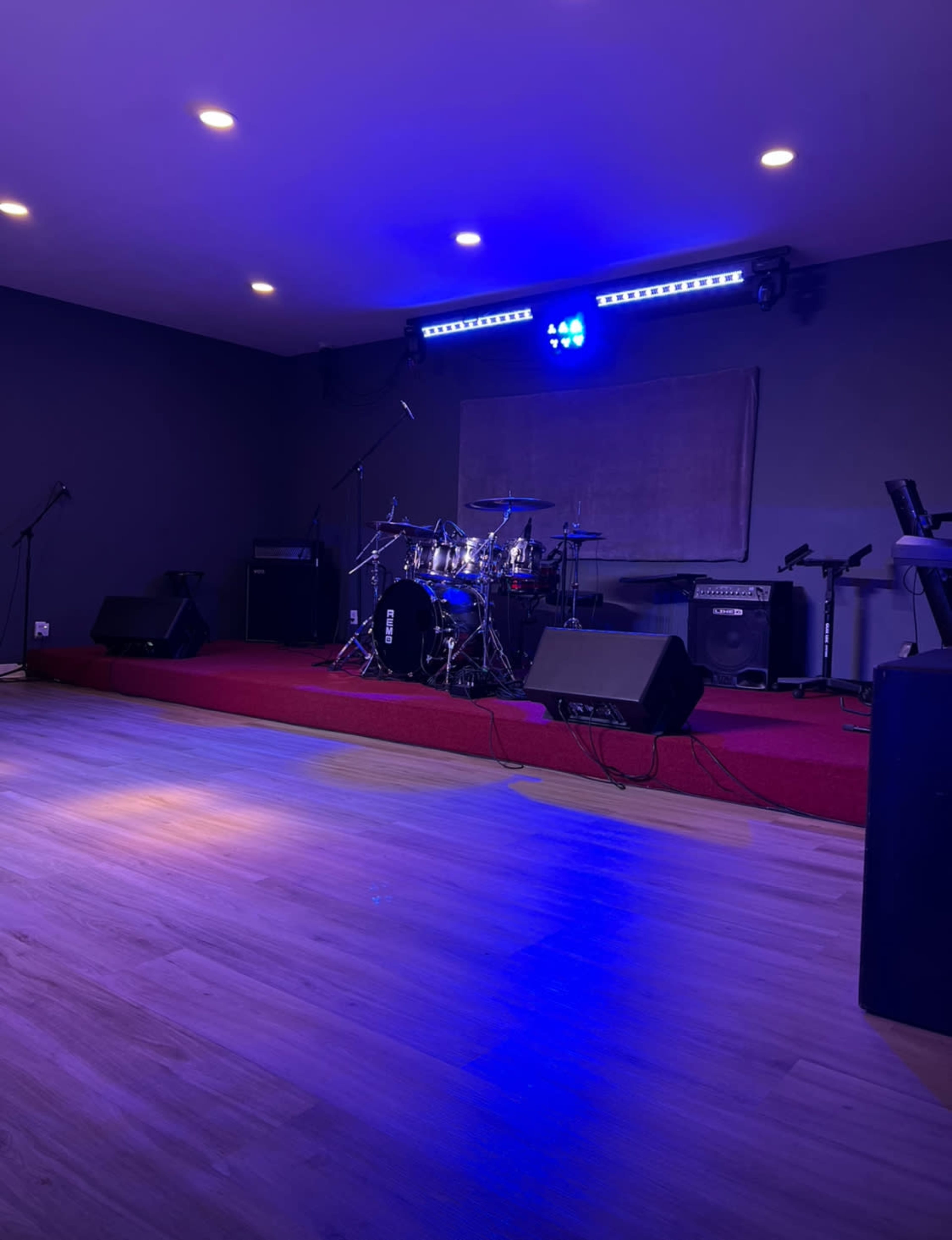 A stage is set up with a drum kit, microphones, and amplifiers on a wooden floor illuminated by blue lights.