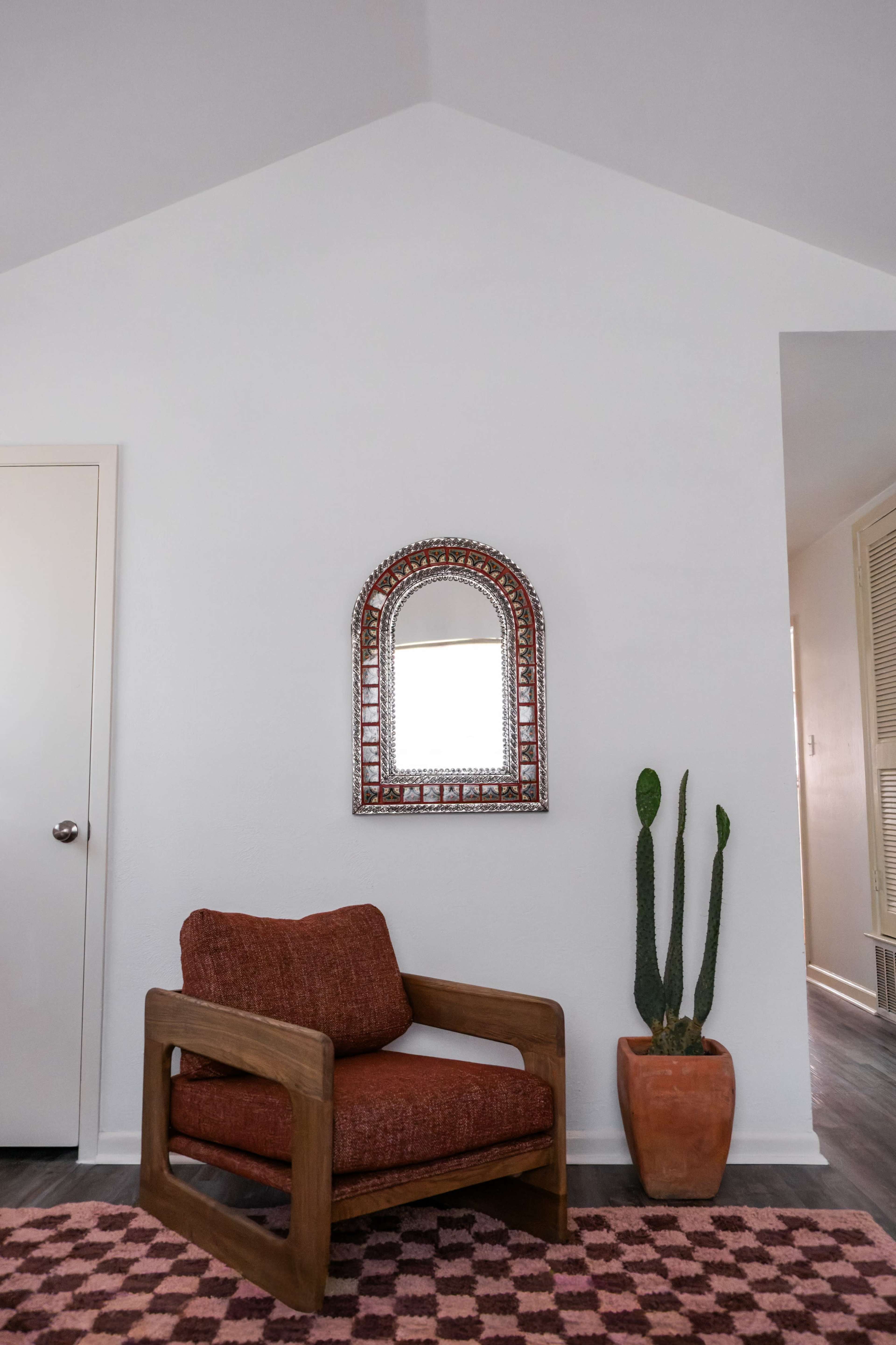 The image shows a cozy living space featuring a wooden chair with a rust-colored cushion, a decorative mirror mounted on the wall, and a potted cactus beside it.