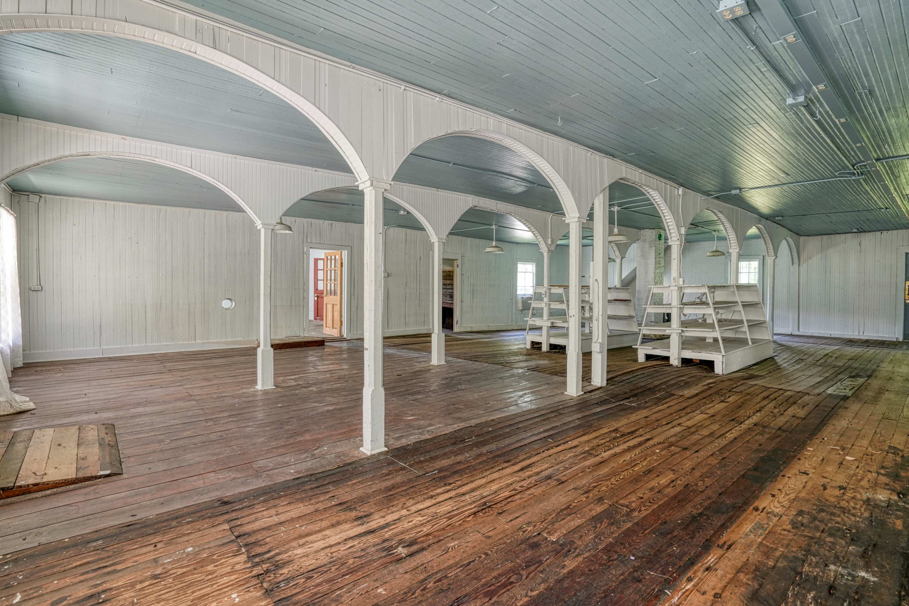 The image shows a spacious, empty room with wooden floors and arched columns supporting a ceiling, featuring several shelves along one wall.