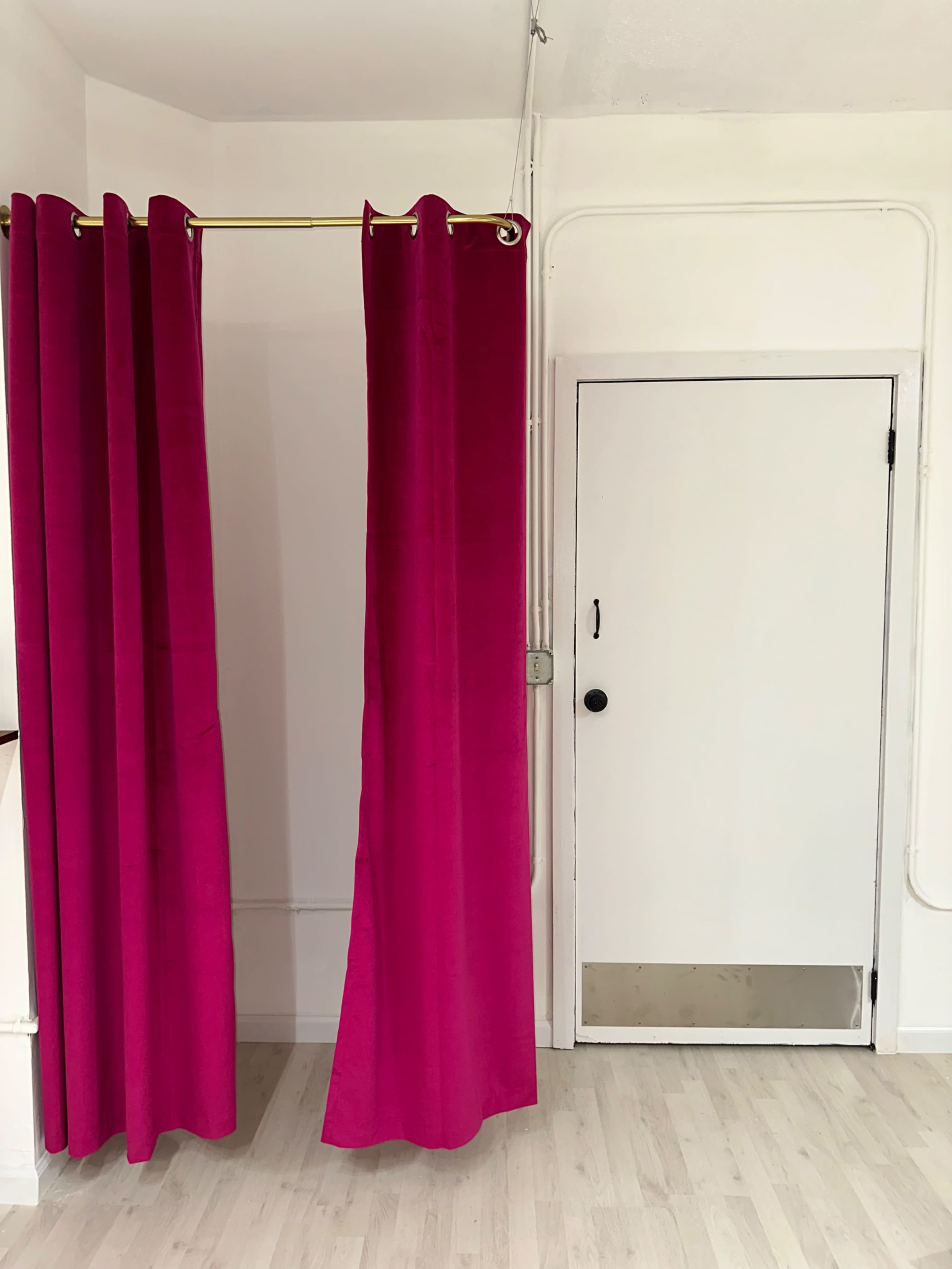 The image shows a pink curtain hanging on a gold rod next to a white door with a black knob in a room with light-colored flooring.