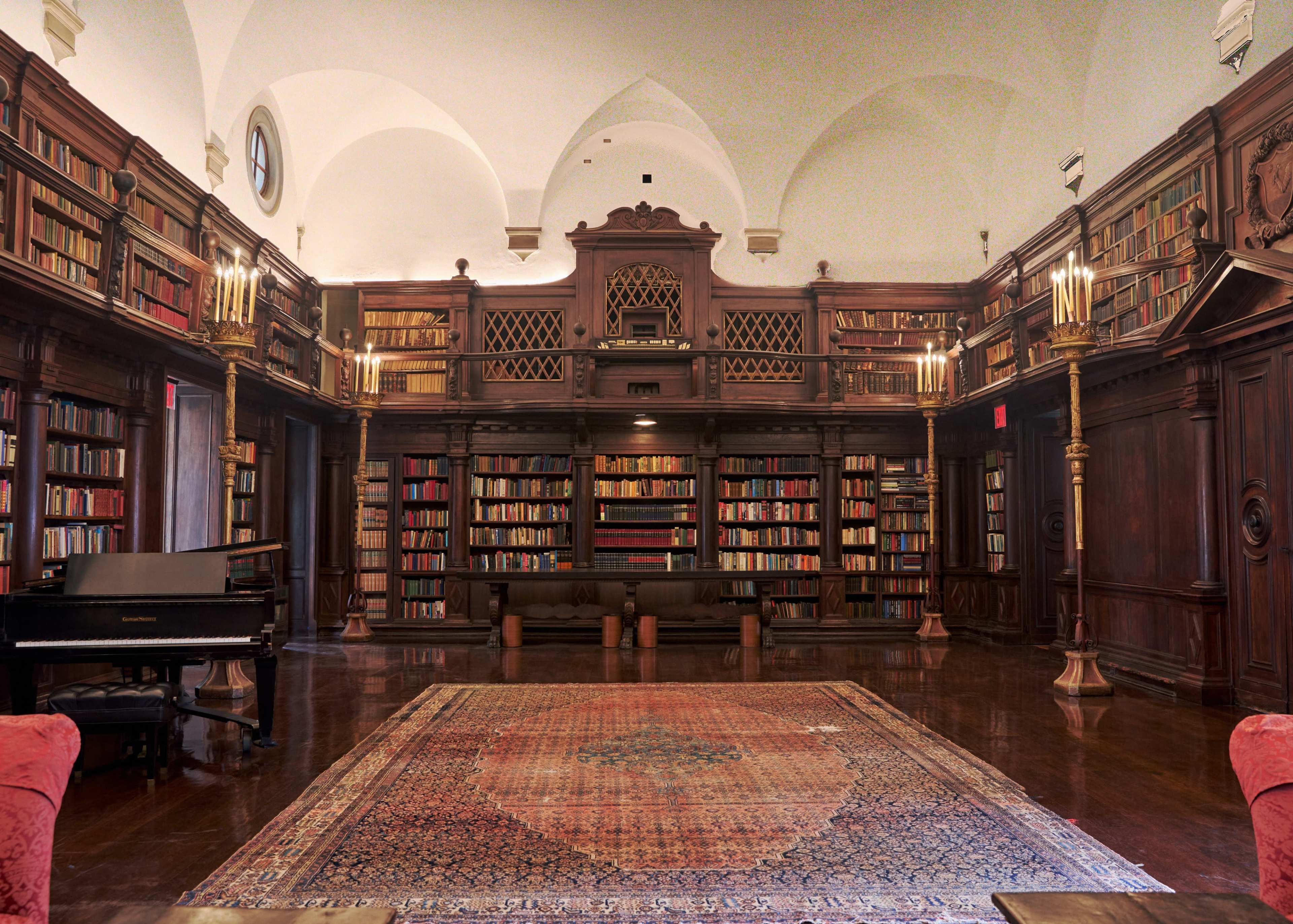 The image shows a dimly lit library with wooden shelves filled with books, a piano in the corner, and a large area rug on the polished floor.